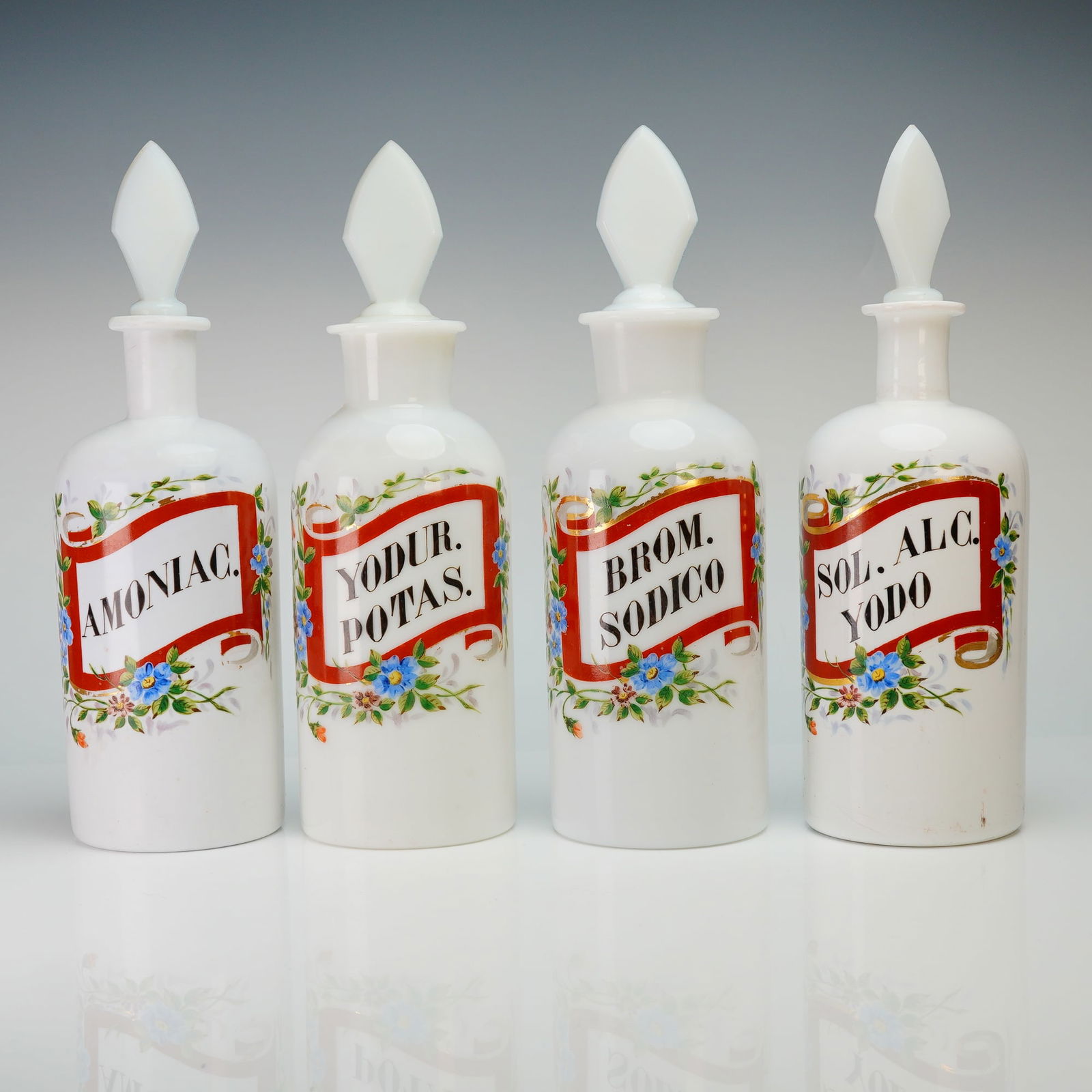 A Set of 4 Milk Glass Apothecary Jars: This 4pc set of milk glass apothecary jars features cylindrical bodies and faceted diamond-shaped stoppers. Each vessel is decorated with hand-painted polychrome floral motifs and red banner labels