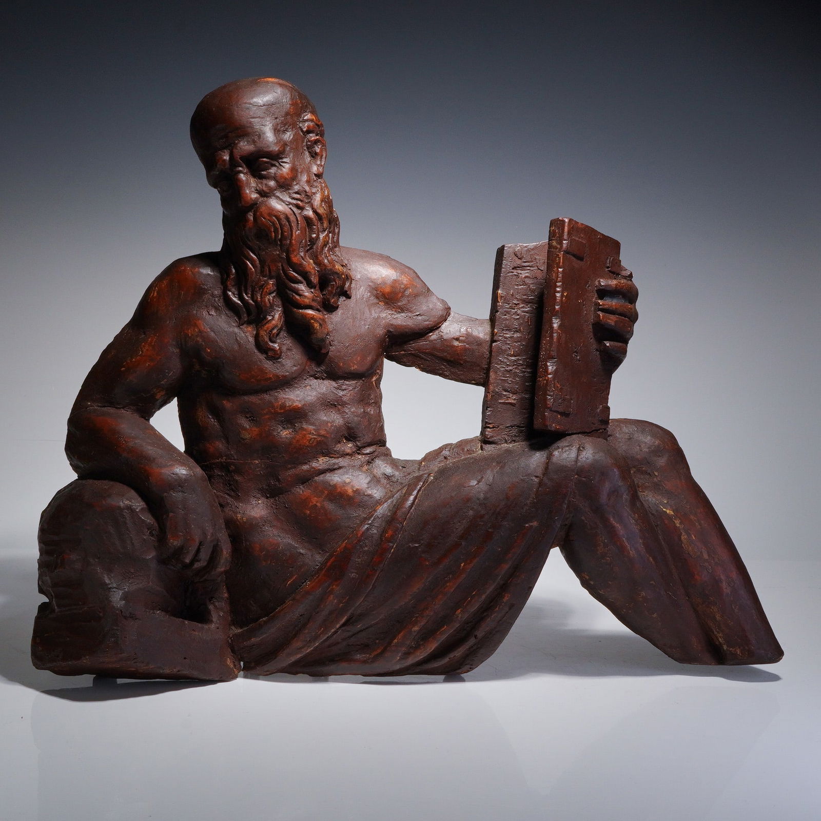Relief-Carved Wood Sculpture of a Philosopher: This relief-carved wood sculpture depicts a seated bearded male figure with a bare torso and draped lower garment, holding a book. The work is executed in a classical manner suggestive of a