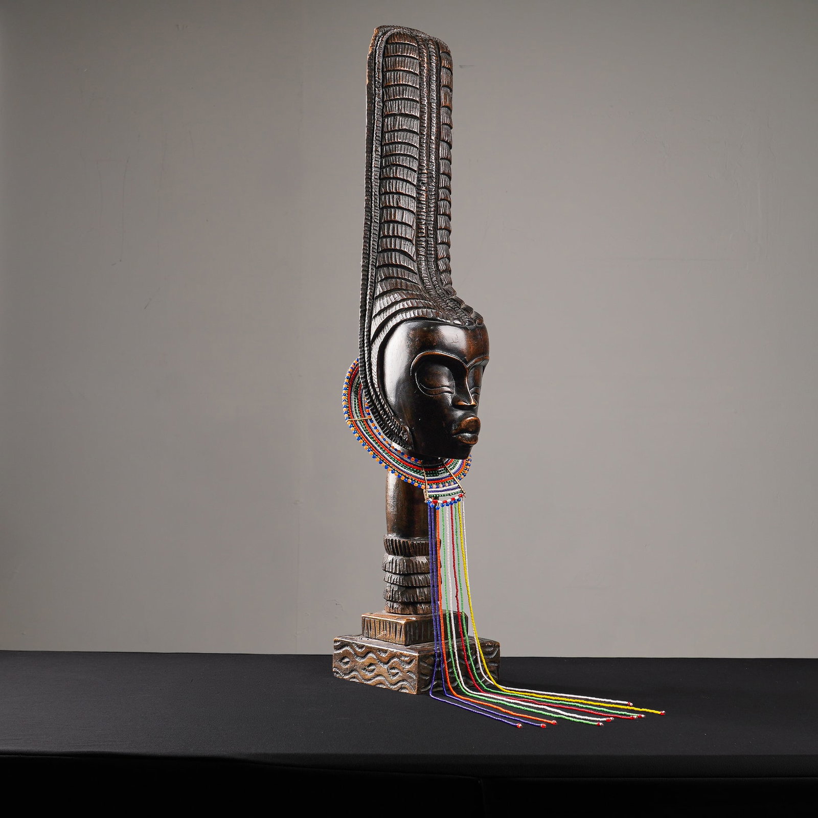 African Baule Wood and Beaded Sculpture of a Woman: This wooden sculpture depicts a woman in beaded adornment. The figure is characterized by an exceptionally elongated, coiffure featuring ridged patterns. The facial features include prominent features