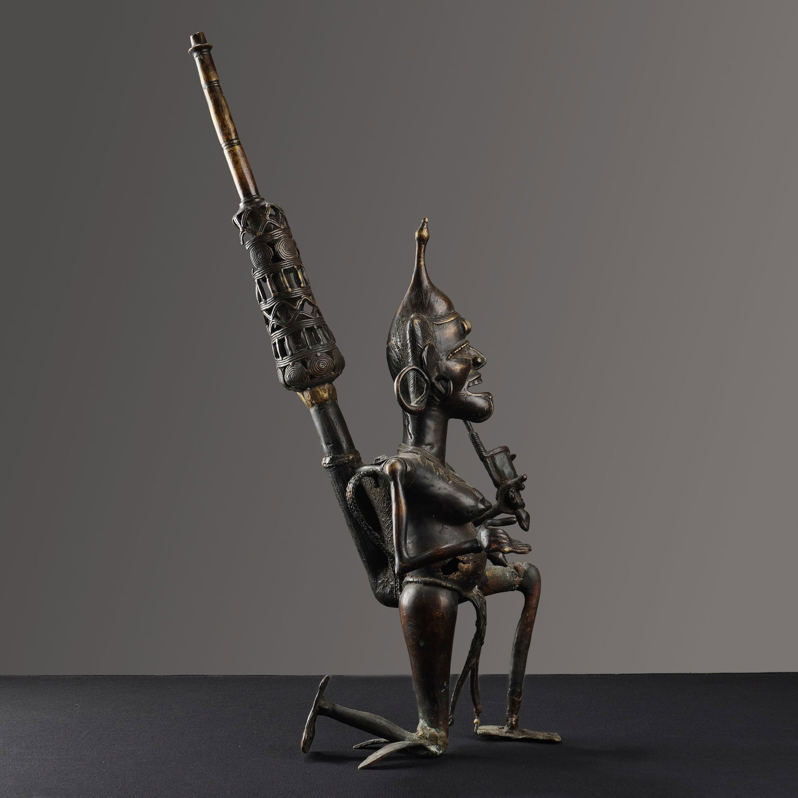 African Bronze Ceremonial Smoking Pipe Sculpture: This bronze sculpture is cast in the form of a ceremonial smoking pipe. The central kneeling male figure is depicted with exaggerated features, including a pointed headdress, large hoop earrings, and