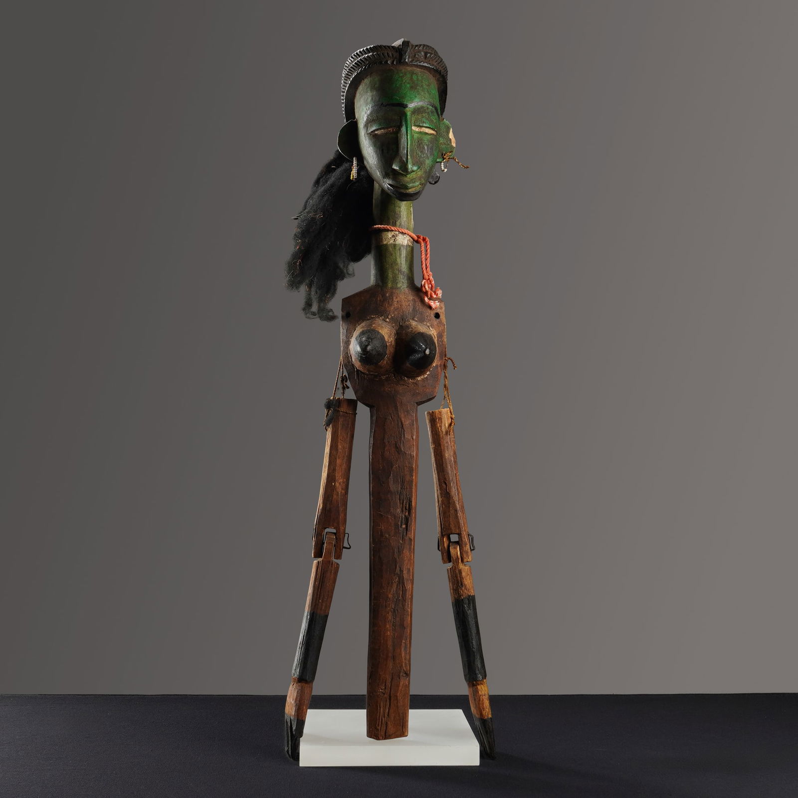 West African Articulated Wooden Female Puppet Figure: This carved wooden female puppet features a green-pigmented head with a carved crest and a mass of black fiber hair. The figure is adorned with small beaded hoop earrings and an orange cord tied aroun