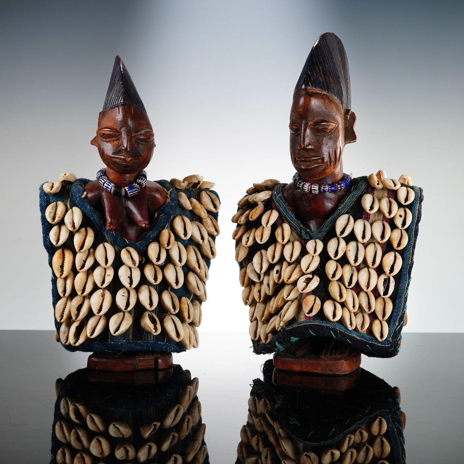 Pair of Yoruba Ere Ibeji Twin Figures with Cowrie Shell Jackets: This pair of African sacred wooden carved figures represents a male and female Yoruba Ere Ibeji from Nigeria. The figures are carved from wood with a dark brown patina and feature tall, textured coiff