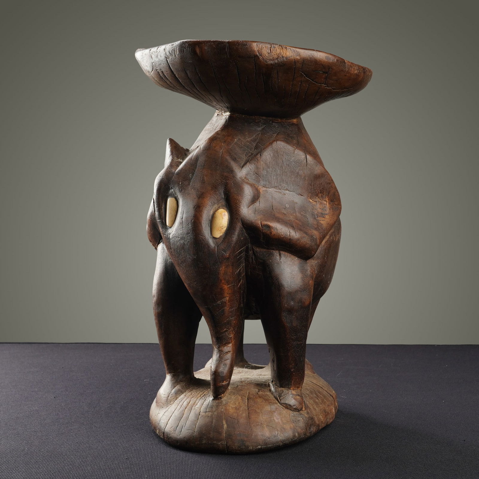 African Carved Wooden Elephant Stool: This African carved wooden elephant stool is rendered from a single block of dark-stained hardwood. The piece features a stylized elephant figure with large ears and a lowered trunk, acting as the ped