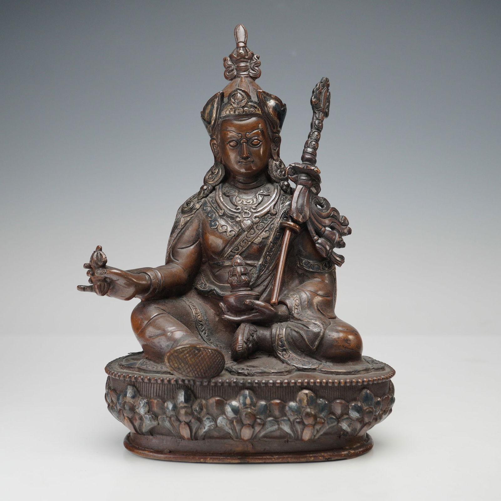 Tibetan Bronze Seated Deity with Silver Overlay: A Tibetan bronze figure of a seated deity, portrayed in a relaxed posture upon a lotus base, grasping a removable staff in one hand and a ritual object in the other. The figure is richly embellished w