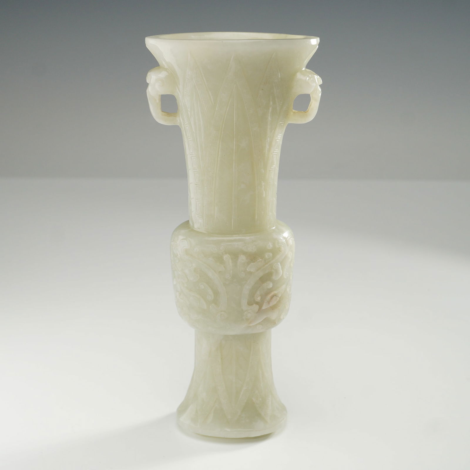 Chinese Carved Nephrite Jade Gu Vase: Featuring a carved nephrite jade vessel adorned with taotie mask and lappet motifs in relief, complemented by finely incised geometric and foliate patterns across the surface. The piece is crafted wit