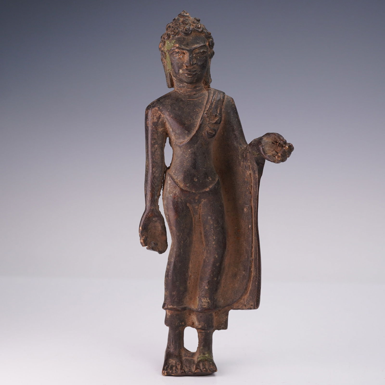 Southeast Asian Bronze Buddha Figurine: Southeast Asian cast bronze figurine depicting the Buddha standing with the right arm extended downward and the left arm bent at the elbow. The figure is shown wearing a long robe draped over the left