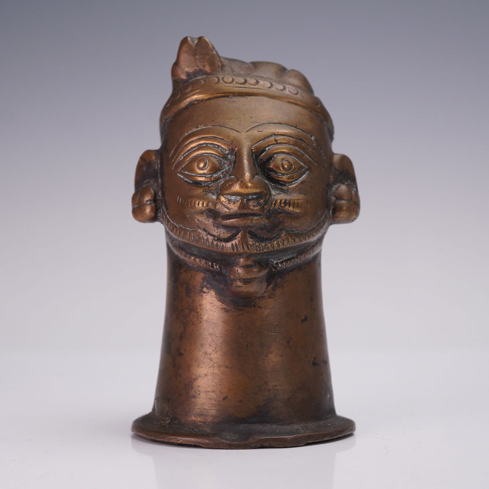 Indian Bronze Male Head Figure: Small Indian bronze bust depicting the head of a man, cast in a cylindrical form with a flared base. The figure features incised detailing to the facial features, including almond-shaped eyes, a musta