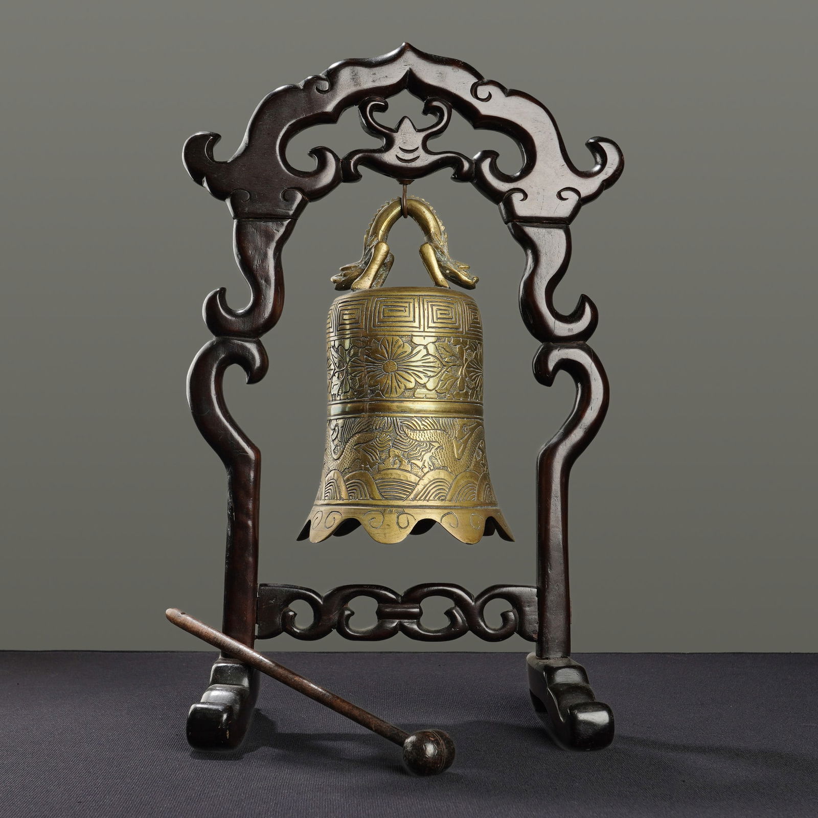 Chinese Brass Temple Bell on Carved Wooden Stand: This lot features a Chinese brass temple bell suspended within an intricately carved dark wood frame and accompanied by a small wooden striker. The cast bell is topped with a double-headed dragon