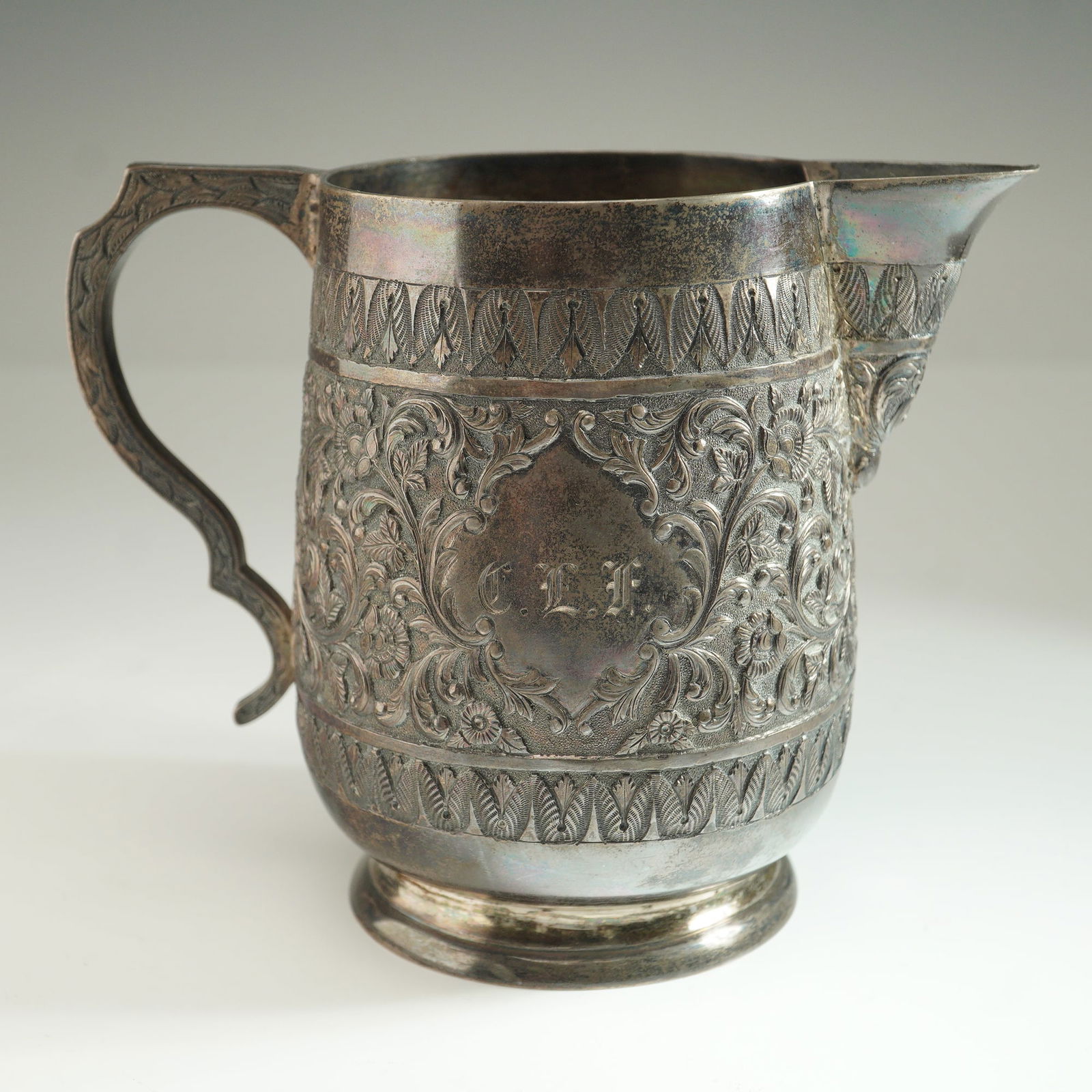 Indian Silver Repousse Creamer: An Indian silver creamer of traditional form, executed with intricate repousse decoration featuring a scrolling floral motif. The handle is detailed with complementary engraved embellishments.