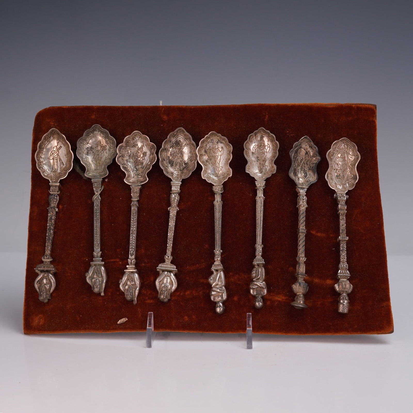 8pc Thai Silver Decorative Spoons: Set of eight Thai silver spoons displayed on a velvet board, each featuring an intricately cast handle topped with a detailed figure and a scalloped bowl engraved with traditional scenes and motifs.