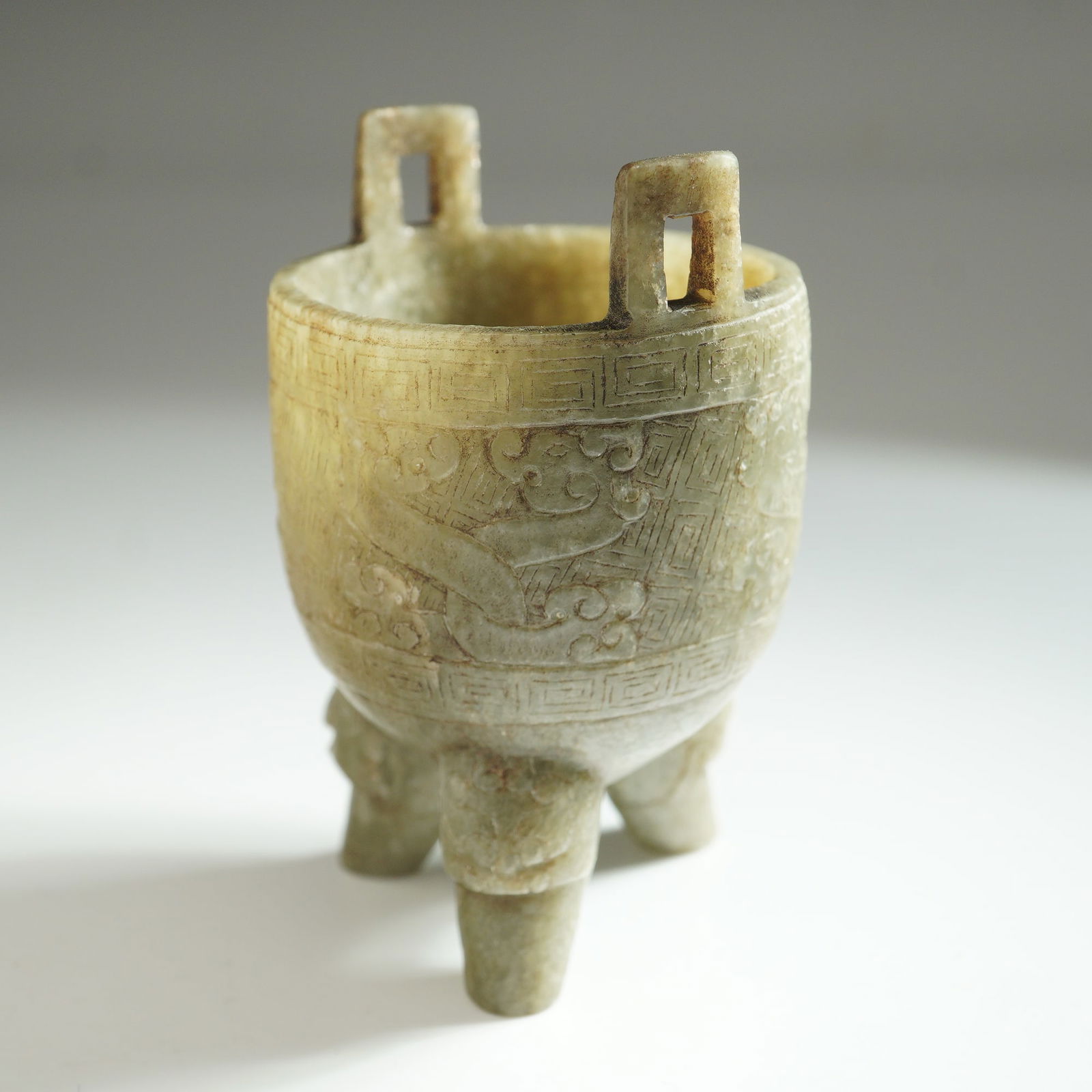 Chinese Ming Jade Archaic-Style Tripod Vessel: Small tripod vessel carved from pale green stone. The vessel is supported on three sturdy legs and features two rectangular handles extending vertically from the rim. The exterior surface is