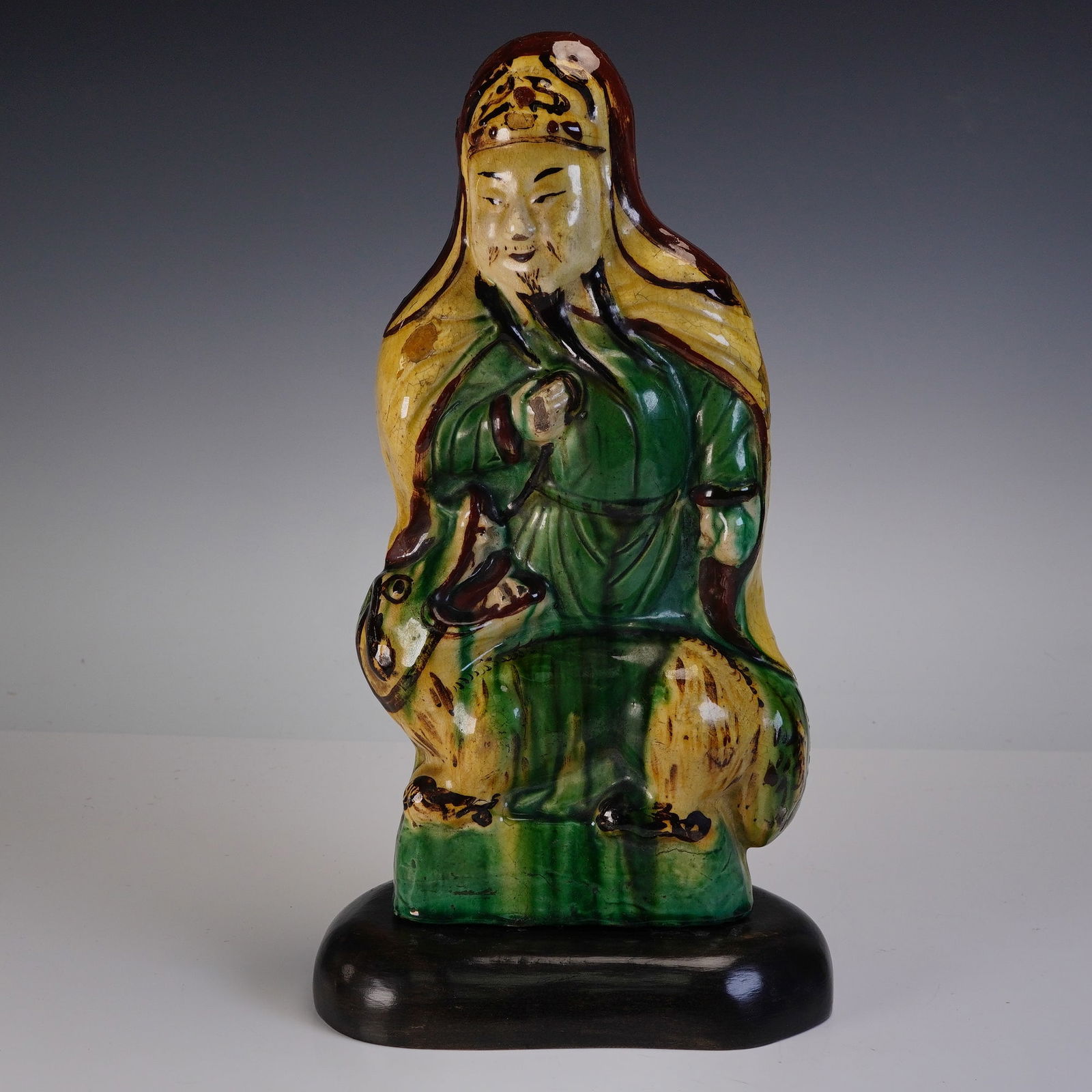 2pc Chinese Ming Pottery Glazed Figure with Wooden Base: The figure is rendered in a standing position with a long robe glazed in green, yellow, and brown tones, and features a bearded male figure with a headdress, holding an object in one hand. At his side