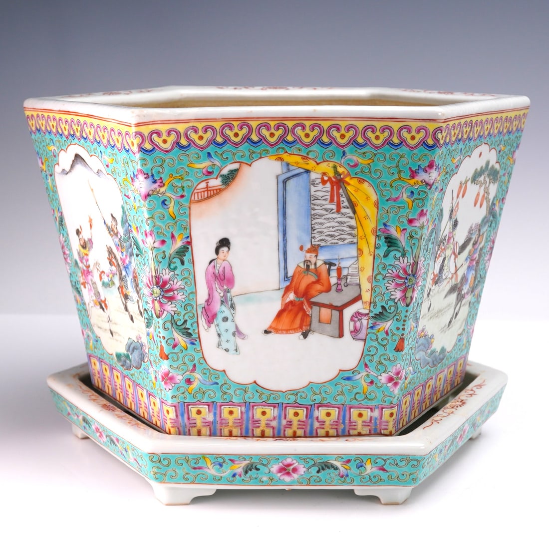 2pc Chinese Guangxu Famille Rose Hexagonal Planter with Underplate: The porcelain planter features six panels, each decorated with hand-painted figural and landscape scenes, including court figures, warriors on horseback, and women in garden settings. The panels are b