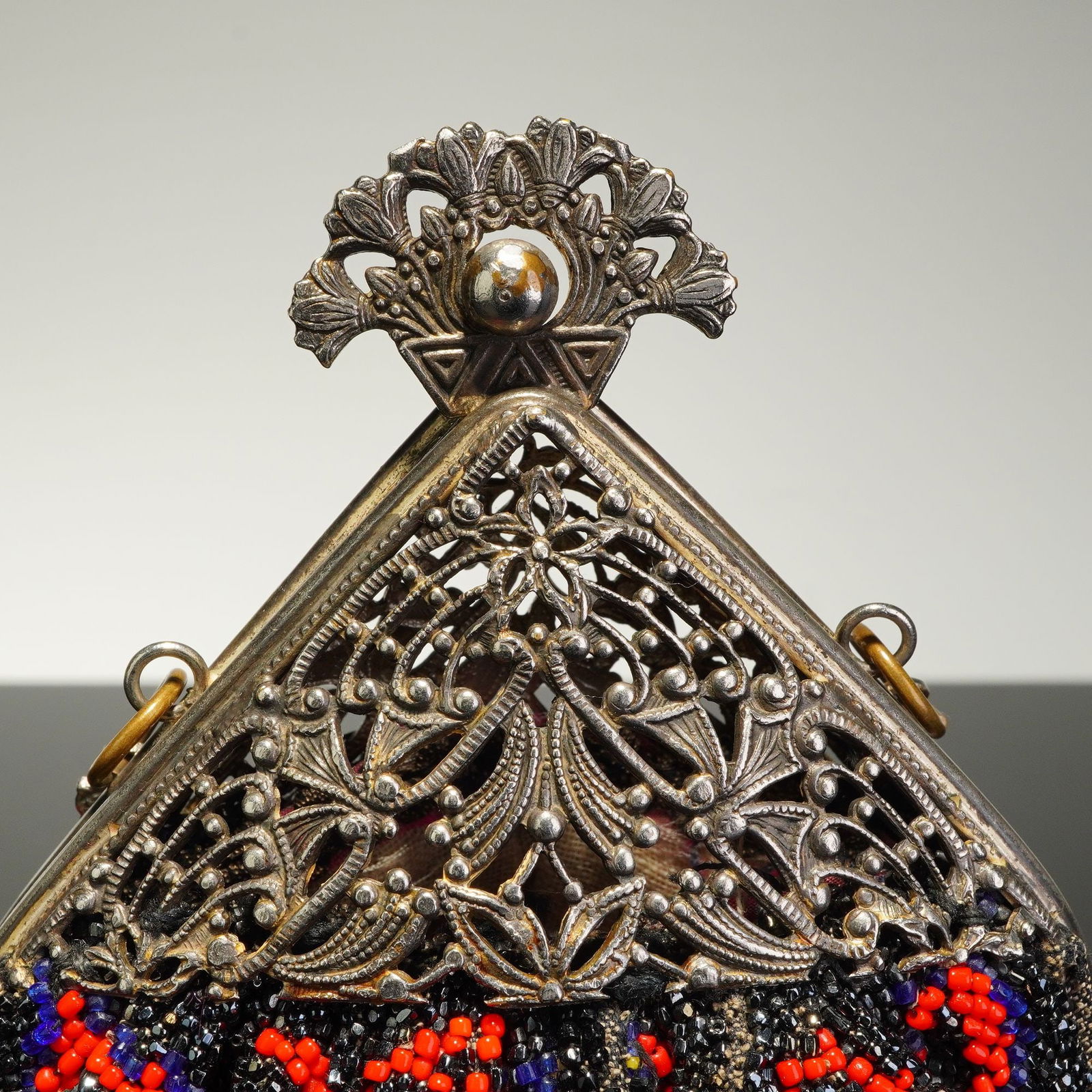 Micro Beaded Handbag with Ornate Silver-Toned Filigree Frame: This micro beaded handbag features an intricate floral design worked in red, blue, yellow, and green seed beads against a black ground. The bag is mounted to an ornate triangular silver-tone metal fra