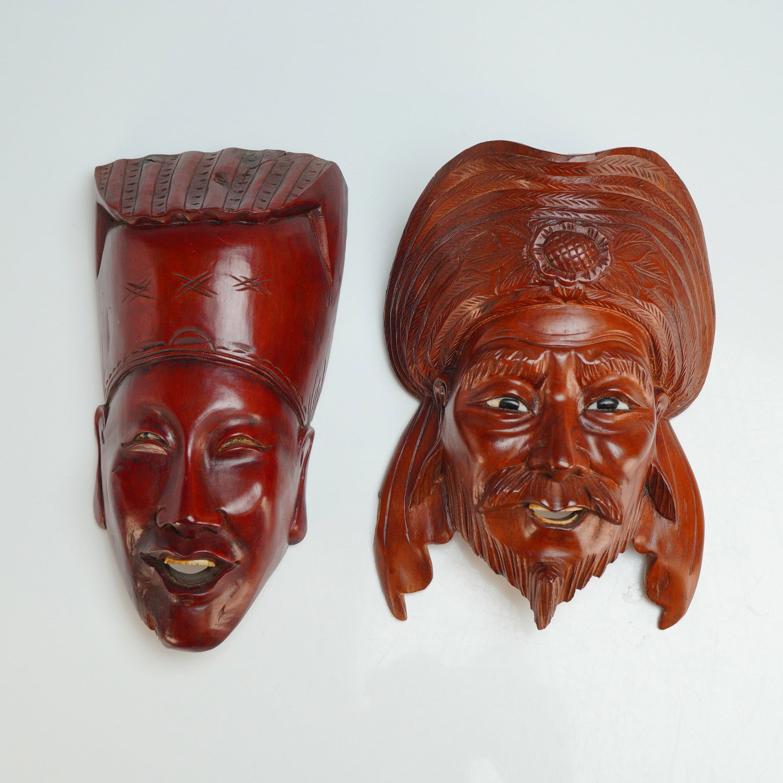 A Pair of Chinese Wooden Wall Masks: A pair of hand-carved Chinese wooden wall masks depicting figures with expressive facial features and elaborate headgear. These pieces are finished in a dark reddish-brown stain and are representative