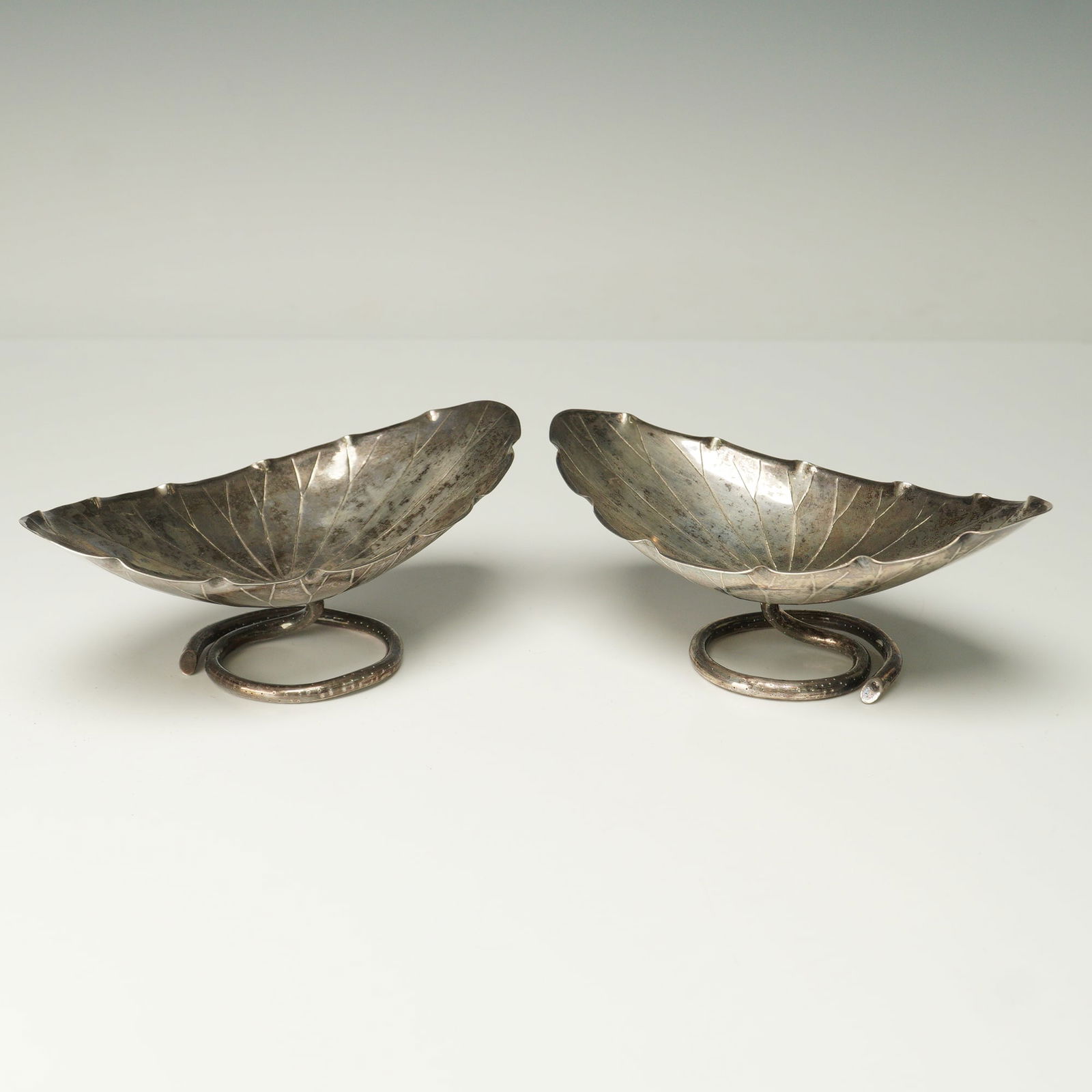 Pair of Japanese Silver Leaf Form Cups on Coiled Stem Bases: This striking pair of silver cups is crafted in the form of naturalistic leaves, their veins and contours meticulously engraved to capture the delicate beauty of nature. The silver has developed a ric