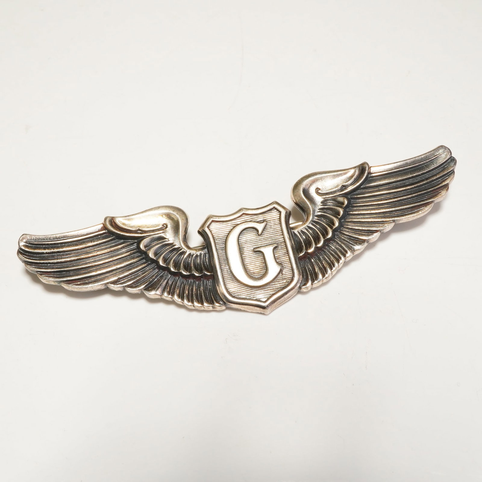 N.S. Meyer Sterling Silver U.S. Army Air Force Glider Pilot Badge: This badge features a central shield embossed with a capital letter G flanked by outstretched feathered wings. The reverse of the insignia is stamped STERLING and includes the shield-shaped hallmark a