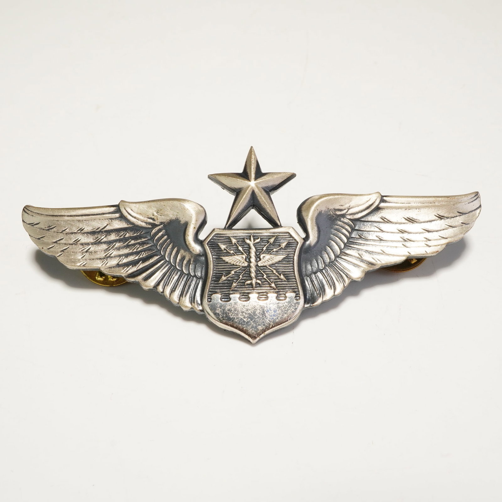 Sterling U.S. Air Force Senior Navigator Wings Badge: The badge features outspread wings flanking a central shield embossed with a wings and arrows motif, surmounted by a single five-pointed star signifying the senior qualification level. The reverse of