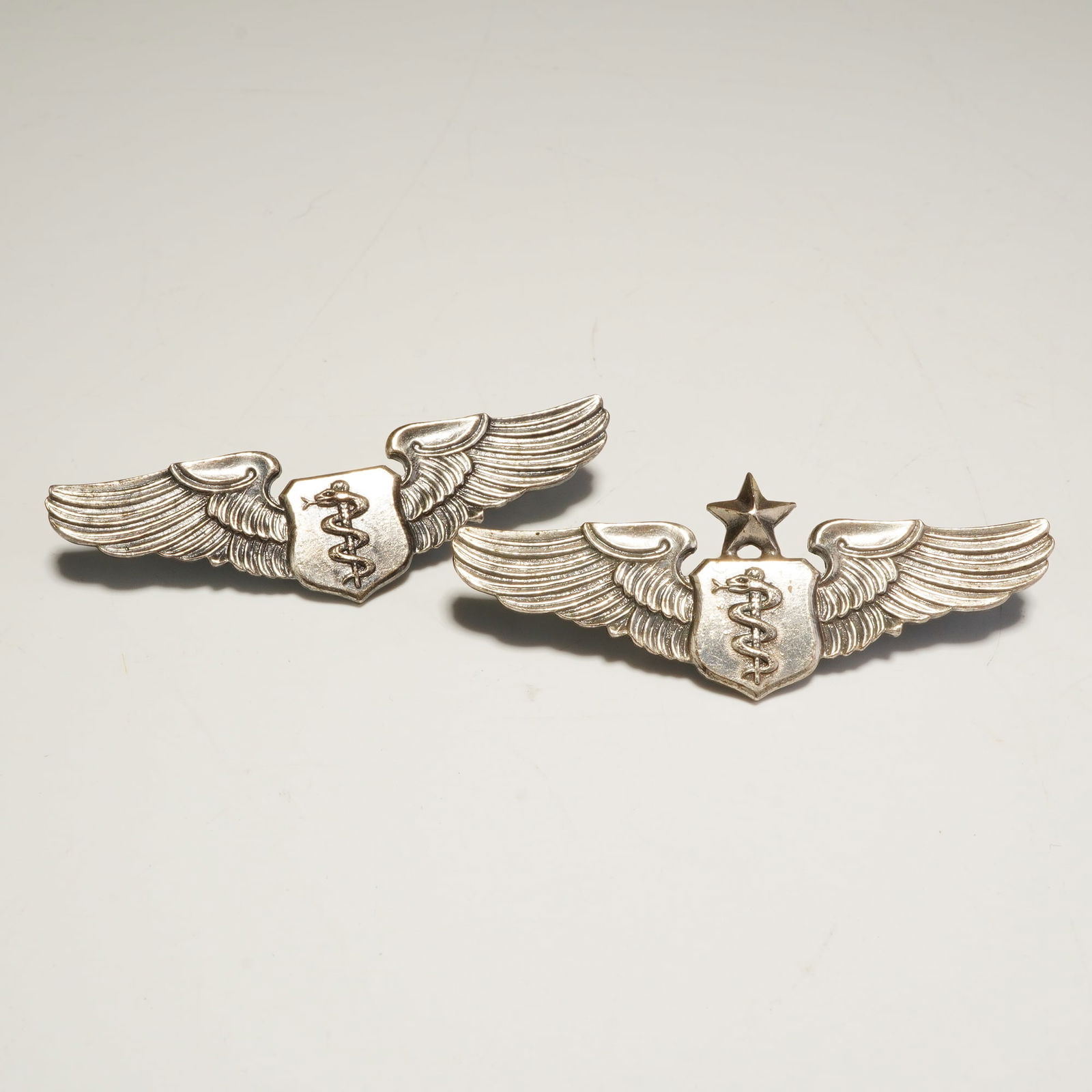 2pc Sterling U.S. Air Force Flight Surgeon Wings: A collection of two United States military flight surgeon insignia badges, including one senior rating example. Each badge is crafted from sterling silver and features outstretched wings flanking a ce