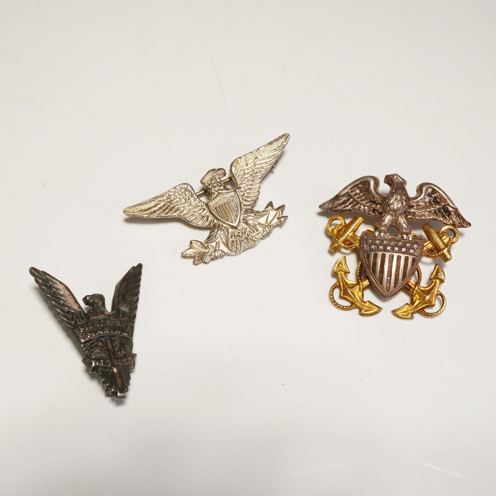 3pc Sterling U.S. Military and Merchant Marine Pins: The lot features a United States Navy officer cap badge with an eagle perched above gold-toned crossed anchors and a central shield. Also included is a U.S. Army Colonel Rank Eagle pin depicting a spr