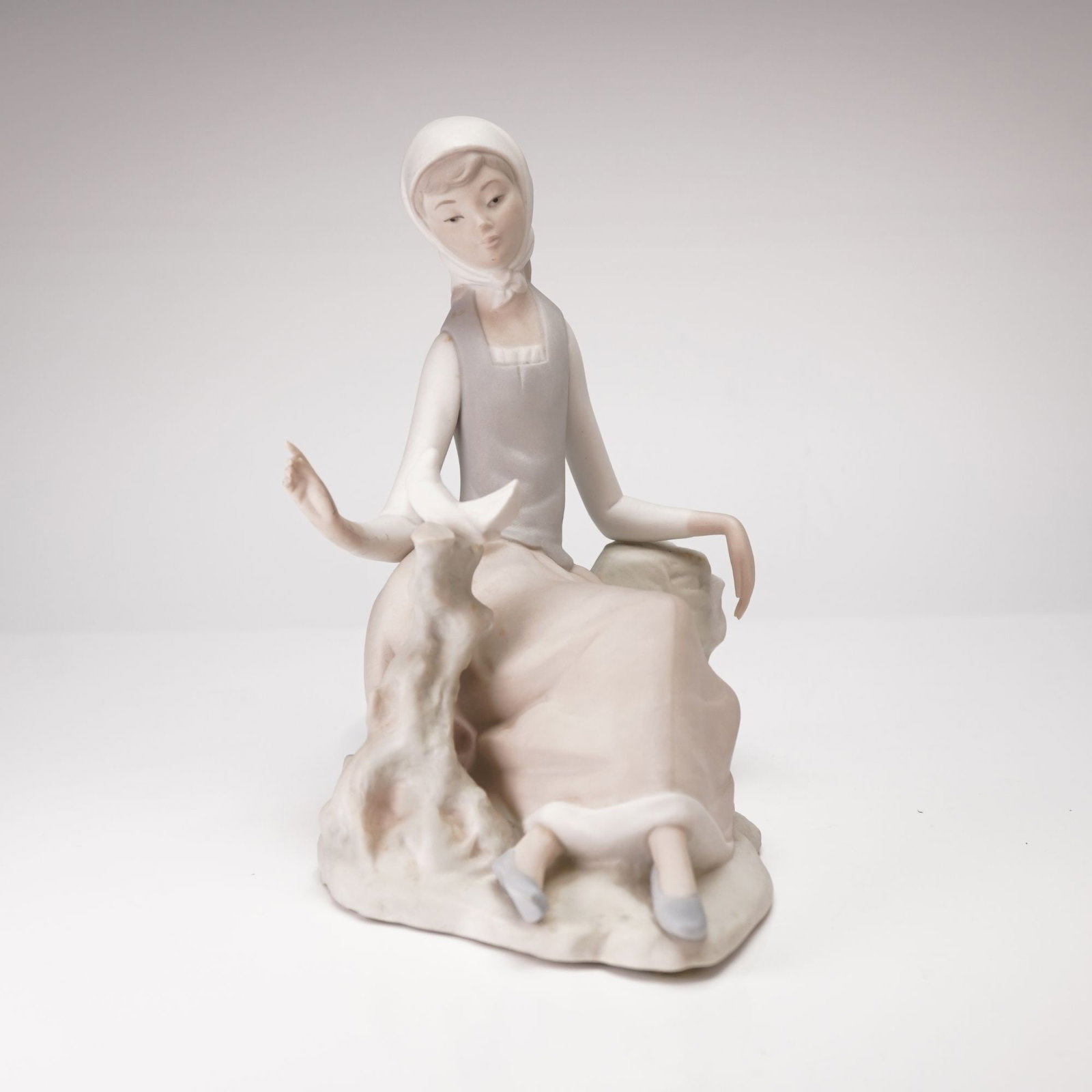 Lladro Figurine, Shepherdess with Dove 1014660: This matte finish porcelain figurine, titled Girl with Goat or Goat's Shepherdess, depicts a young woman in a contemplative pose seated on a textured rocky base. She is accompanied by a small goat res
