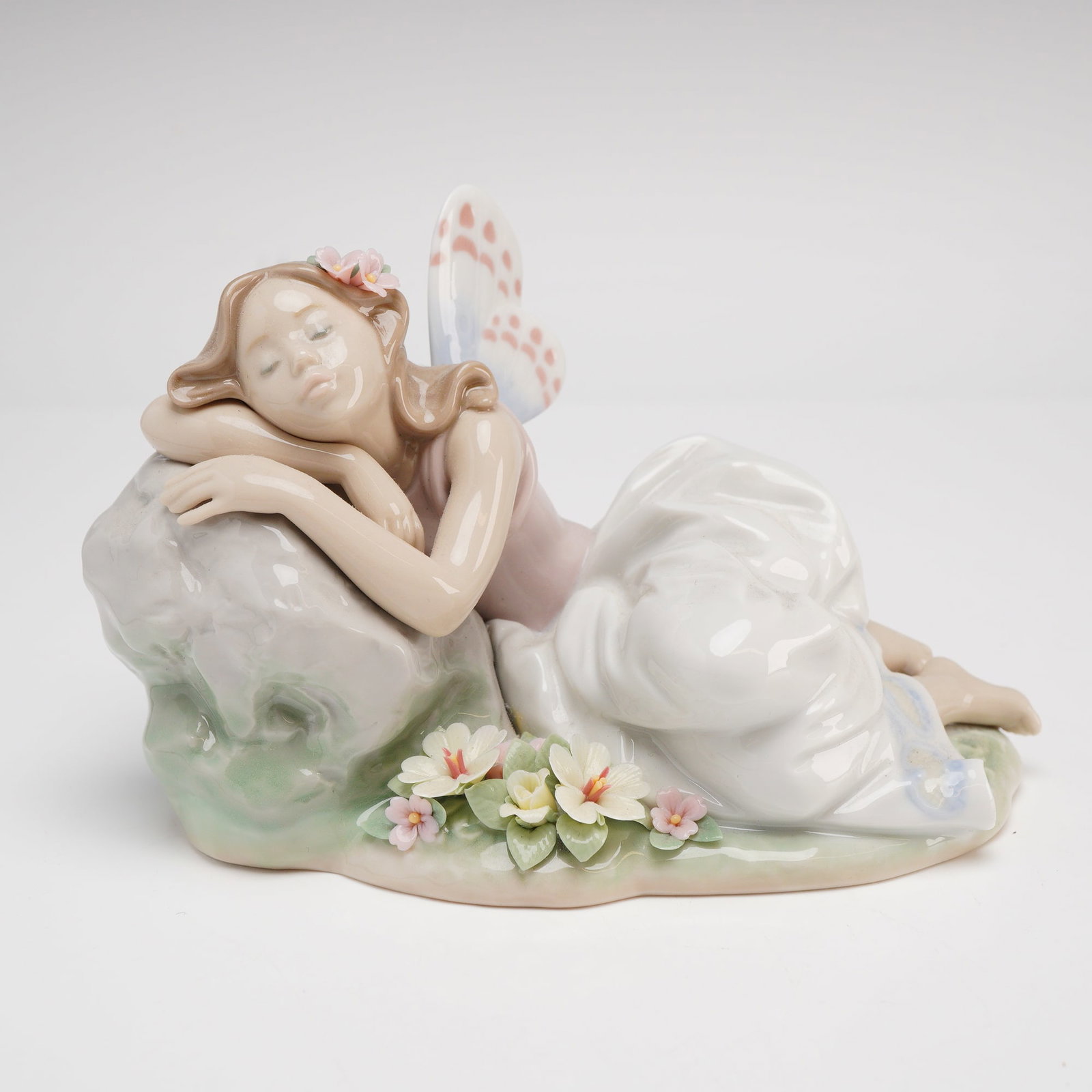 Lladro Privilege Porcelain Figurine, Princess of the Fairies #7694: A fine Lladro Privilege porcelain figurine titled Princess of the Fairies, model number 7694. This piece depicts a reclining winged fairy in a peaceful slumber against a light gray rocky outcrop. The