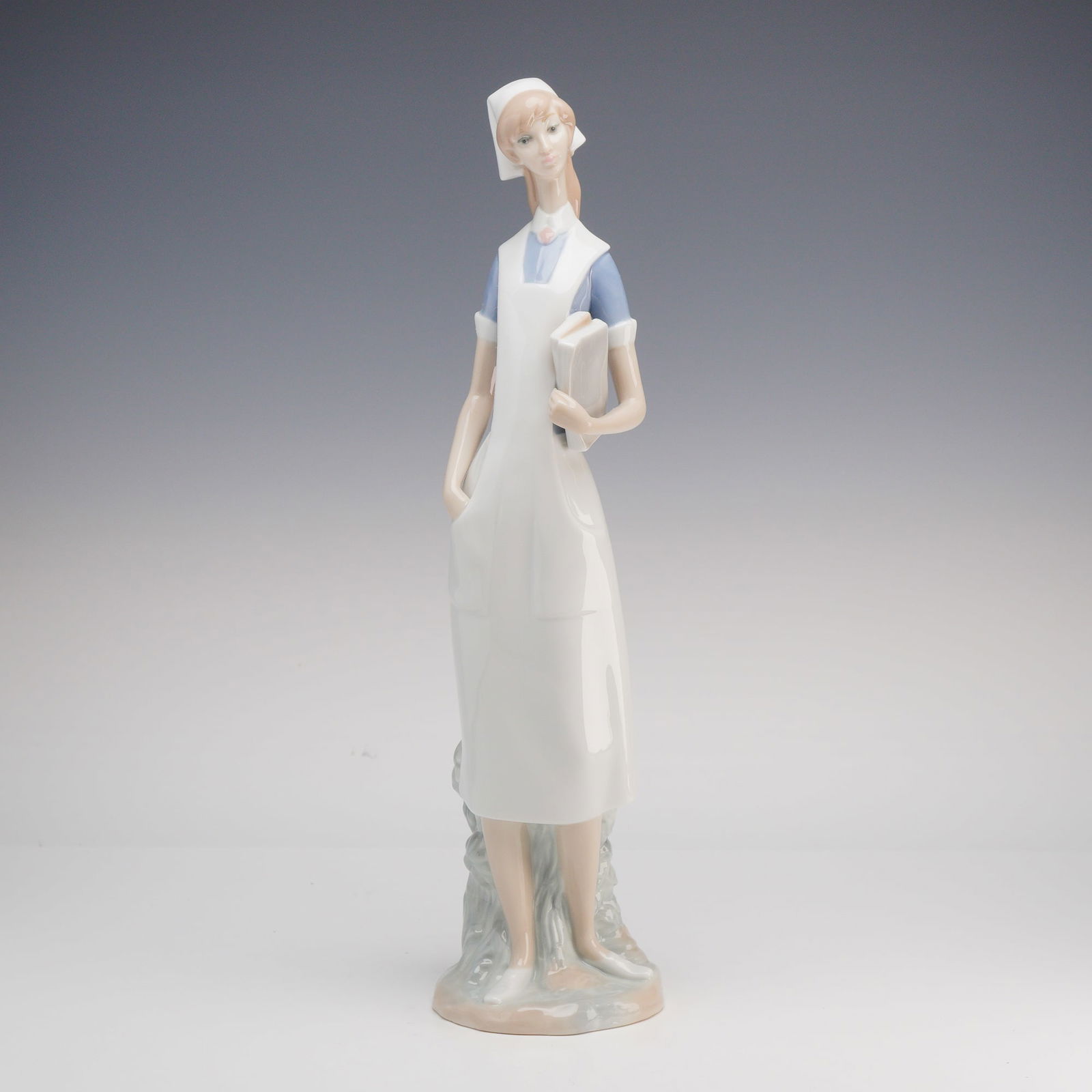 Lladro Porcelain Figurine, Nurse 1004603: Depicting a standing nurse wearing a white cap, blue dress, and white apron, holding a book under one arm with the other hand in the apron pocket. The figure features hand-painted facial details and