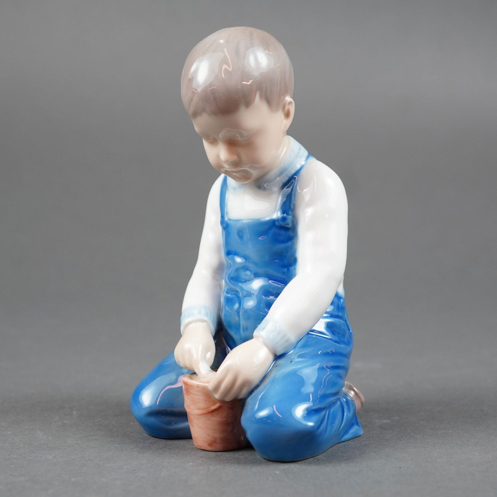 Bing & Grondahl, Boy with Bucket Figurine #2127: A Bing & Grondahl glazed porcelain figurine titled Boy with Bucket, model number 2127. The figure depicts a young boy kneeling on both knees, dressed in blue overalls over a white shirt, focused on a