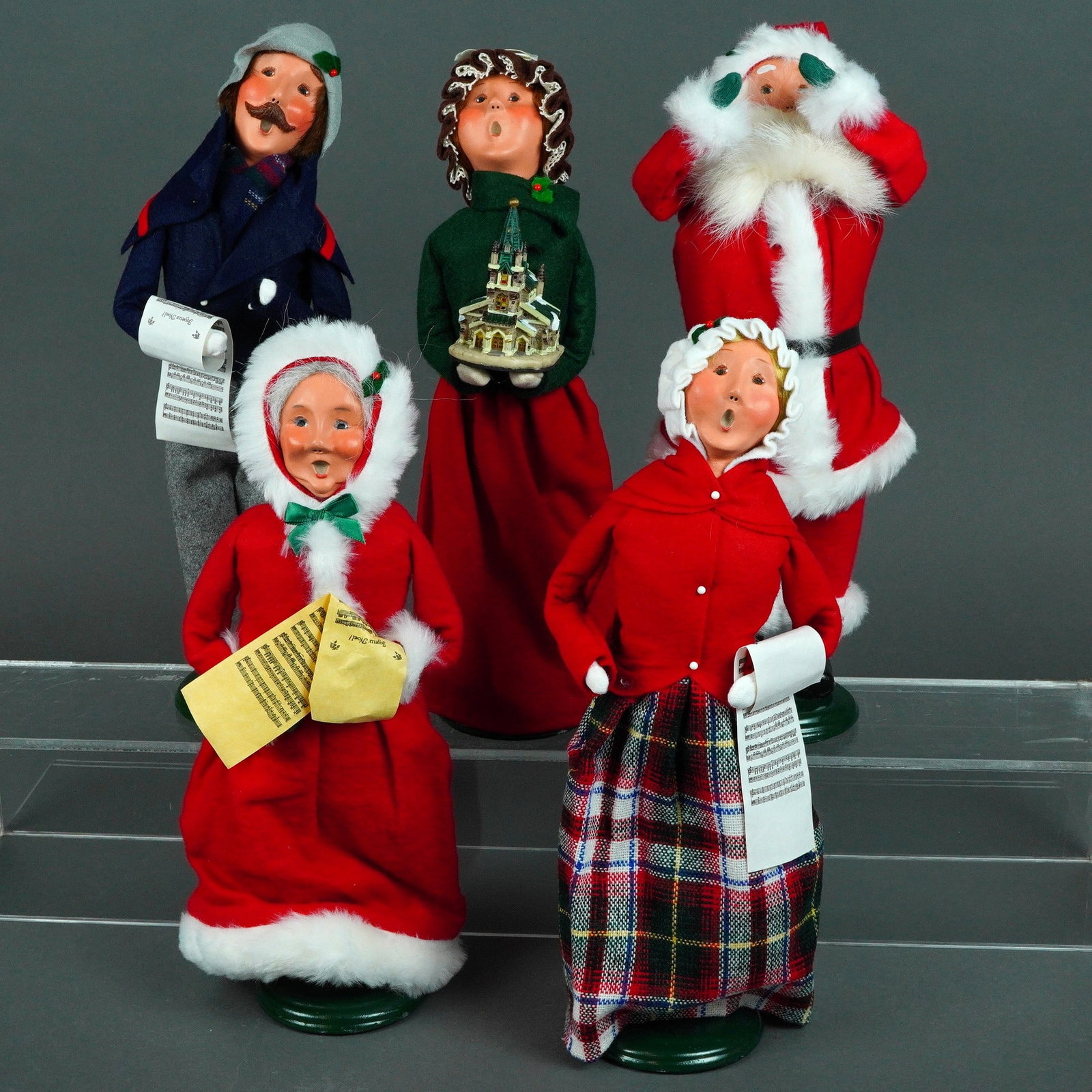 Five Piece Byers Choice Ltd. The Carolers Figurines: Handcrafted figurines of mixed media figures feature modeled porcelain faces with characteristic singing expressions, felted wool, and various fabrics over wire frames. Each figure is mounted on a gre