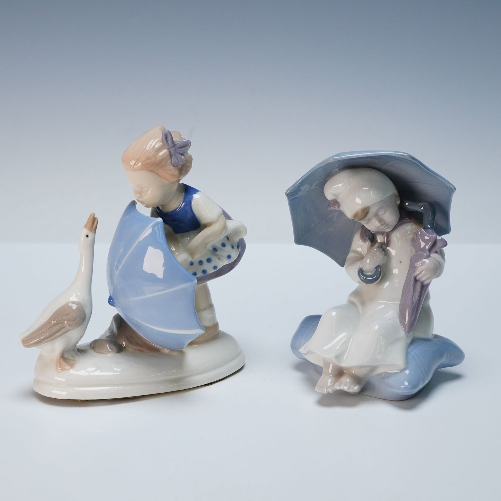 Carl Scheidig Porcelain Figurines - Girl w/ Goose & Seated Child: A pair of Carl Scheidig porcelain figurines, crafted from glazed porcelain in a pastel, stylized manner. The first figurine depicts a fair-haired girl in a blue and white dress, holding a light blue u