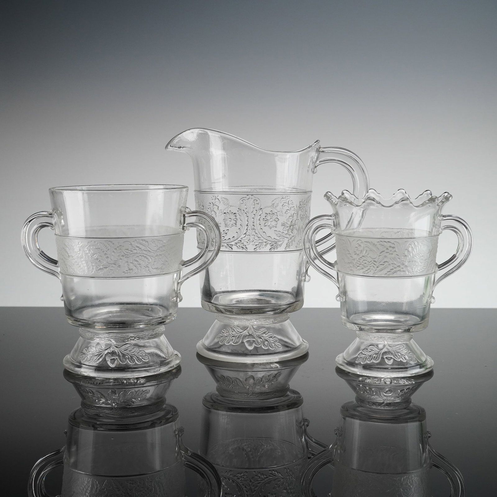 3pc EAPG Glass Pitcher & Dual Handled Vases, Flower Band: This collection of glassware is executed in clear pressed glass. The set consists of three items: a pitcher, a two-handled vase with a ruffled rim, and a matched two-handled vase with a straight rim.