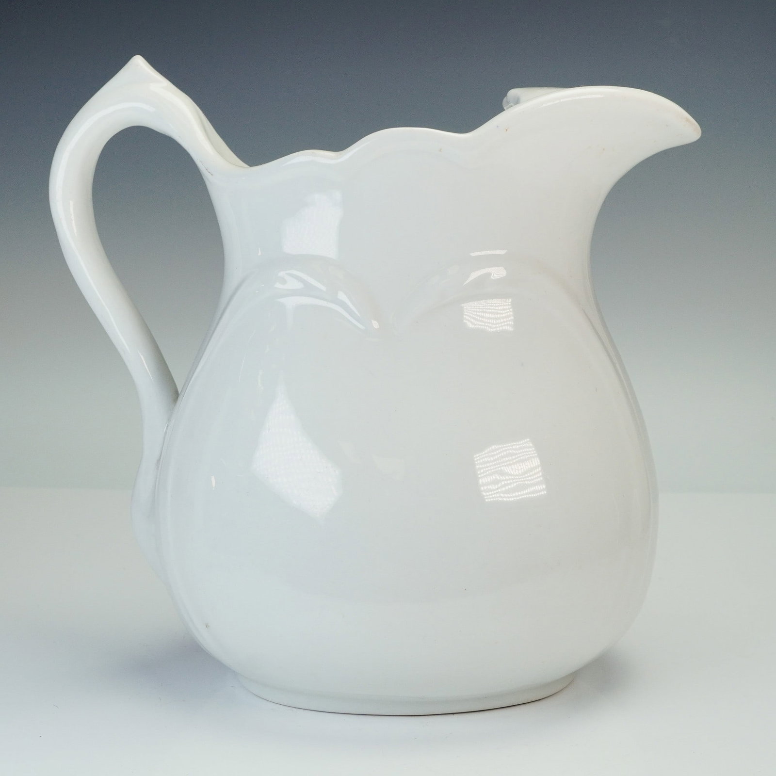 T.P Co. White China Pitcher: A classic white pitcher with a scalloped rim, elegant curved handle, and a distinctive connective piece at the top forming a hole above the spout. Smooth glossy finish and soft rounded body shape