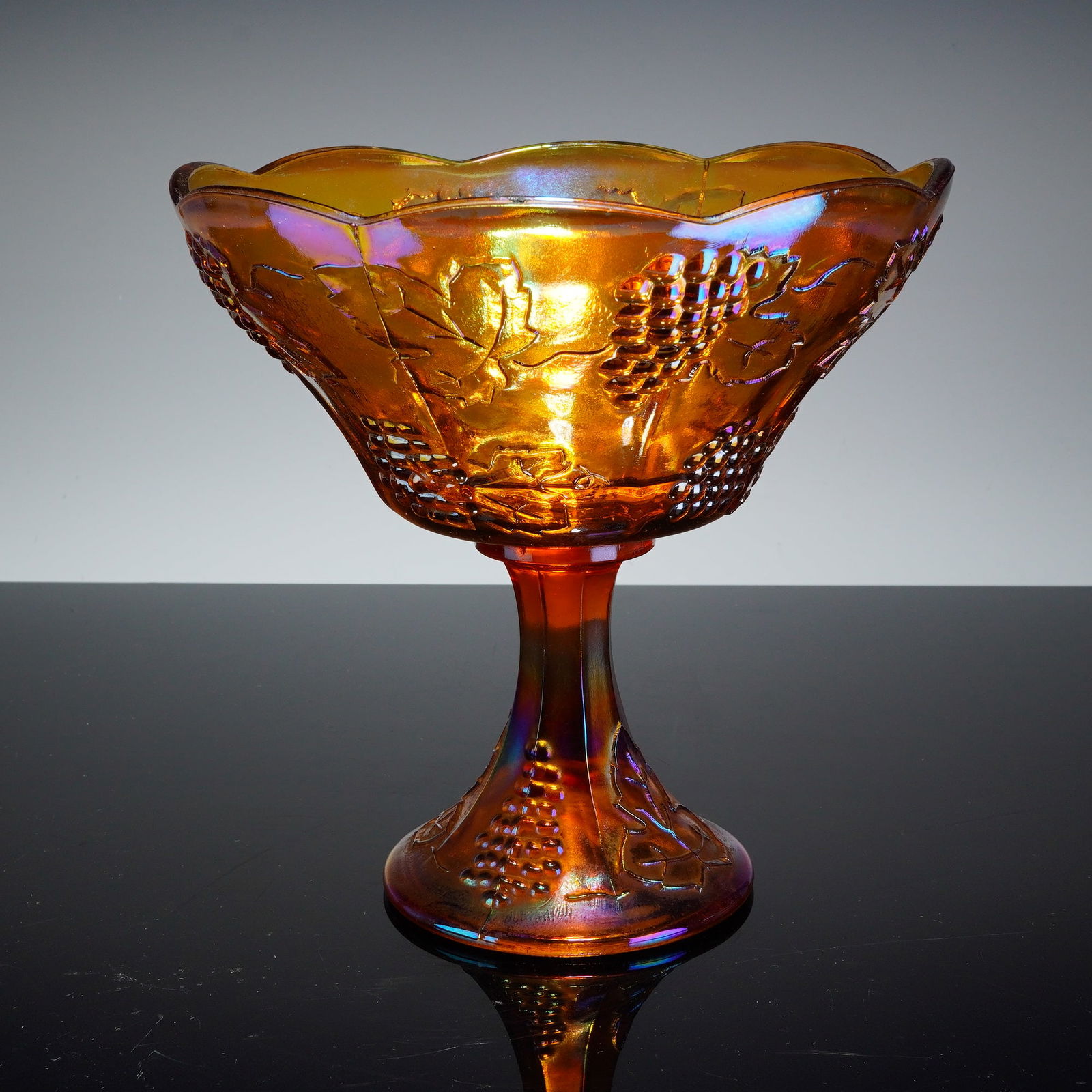 Indiana Glass Iridescent Carnival Glass Pedestal Bowl: This vintage pedestal bowl was produced by Indiana Glass in the Harvest Grape pattern. The piece is a classic example of American carnival glass, featuring a pressed glass construction with a