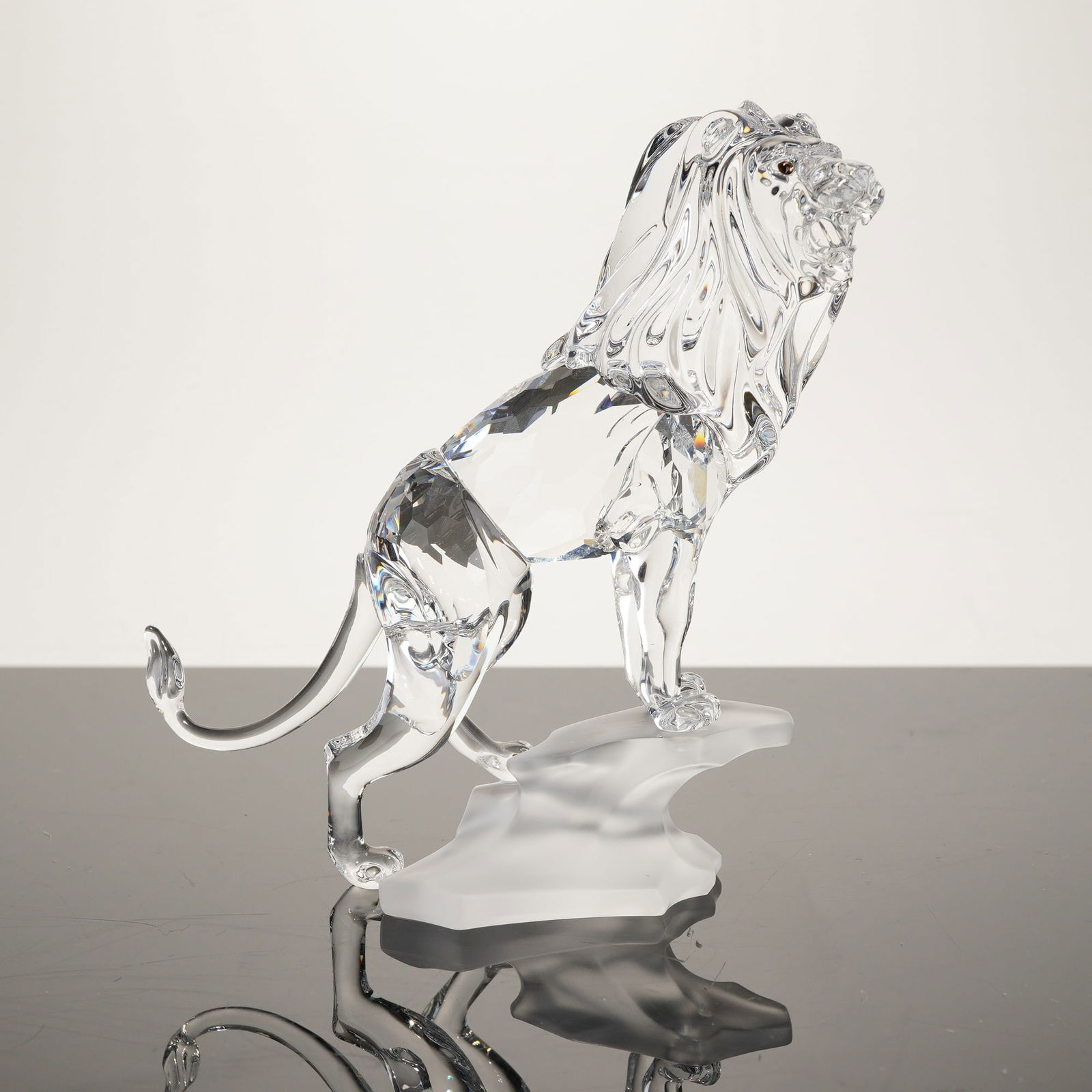 Swarovski Lion Standing on a Rock Figurine #269377: This Swarovski crystal figurine depicts a proud male lion in an alert, standing posture on a frosted crystal base resembling a rocky outcrop. Part of the African Wildlife collection, the piece is craf