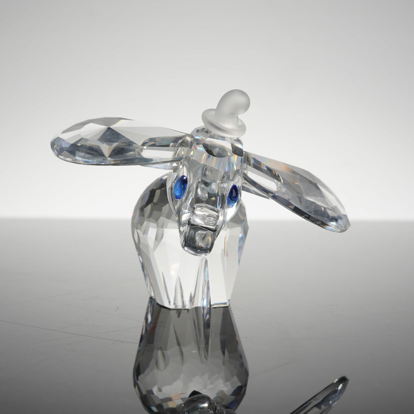 Swarovski Disney Dumbo with Blue Eyes 7640 NR 100 001: A Swarovski crystal figurine depicting the Disney character Dumbo the Flying Elephant. This piece is crafted from clear faceted lead crystal with large outstretched ears. The figurine is distinguished