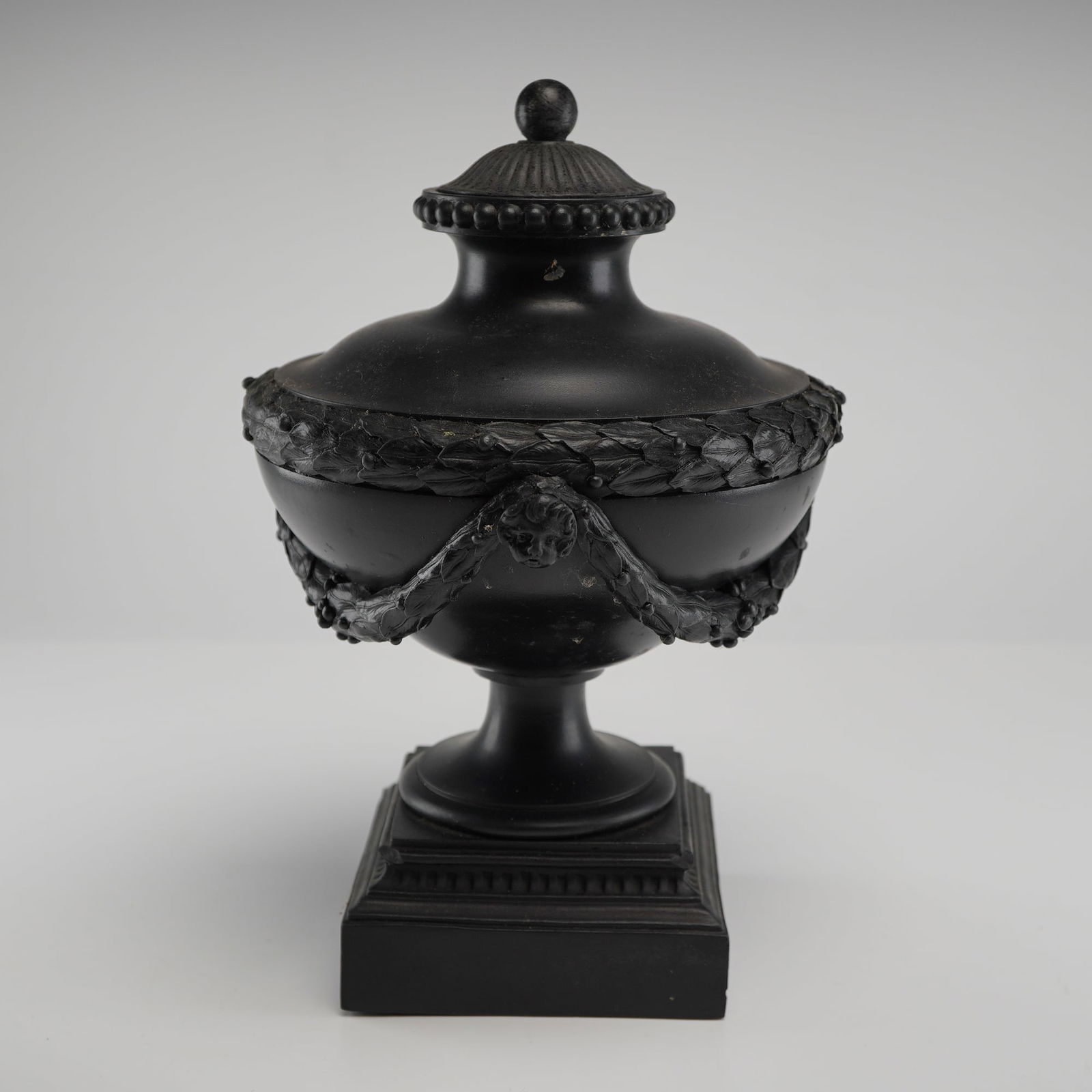 Brass-Mounted Wedgwood and Bentley Black Basalt Lidded Urn: A Neoclassical Wedgwood and Bentley black basalt lidded urn of classical ovoid form. The stoneware body is decorated with sprigged laurel leaf garlands draped from bacchic masks and cherub-head medall