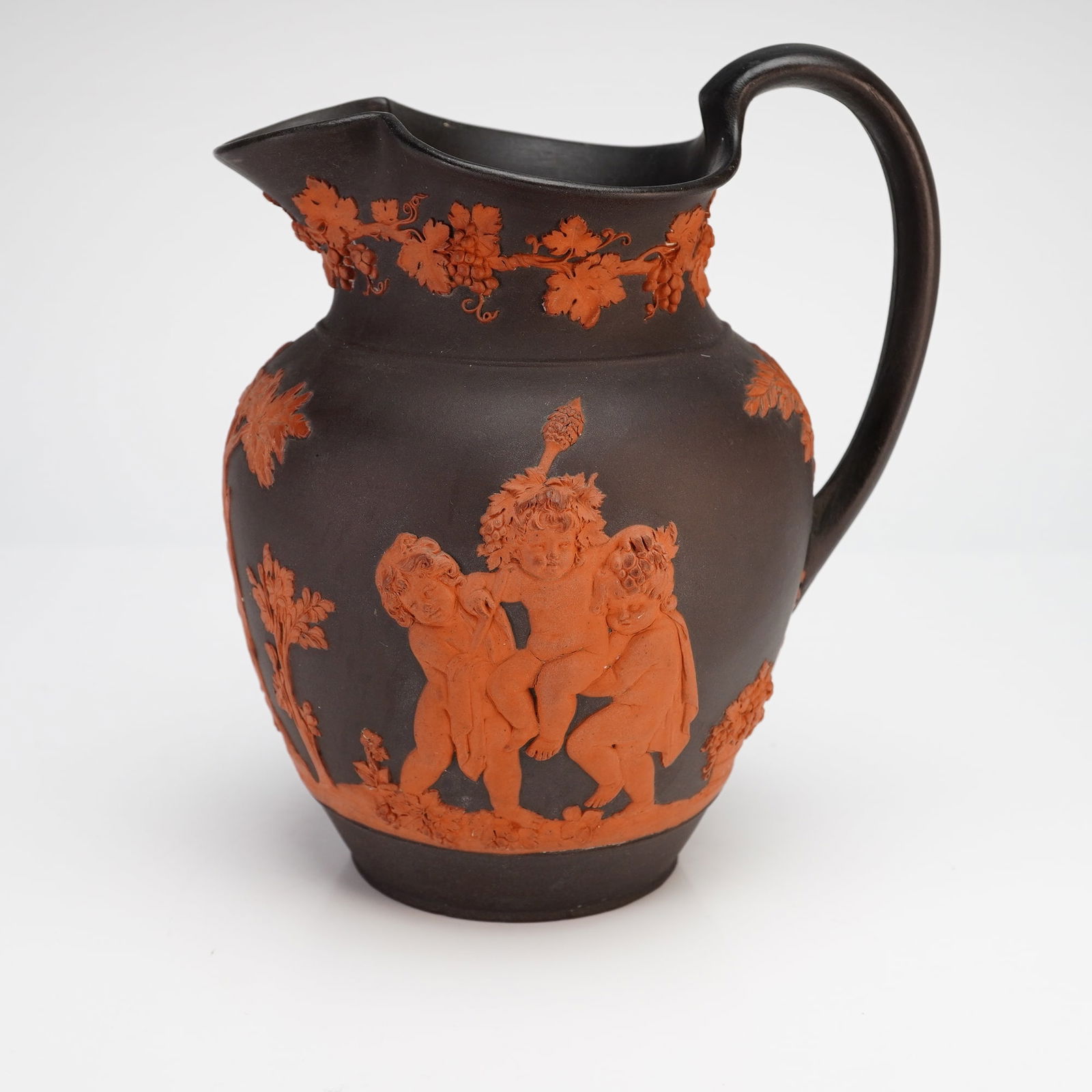 Wedgwood Rosso Antico and Black Basalt Bacchanalian Boys Playing Pitcher: This Wedgwood pottery pitcher is constructed of black basalt with terracotta-colored rosso antico sprigged reliefs in the Bacchanalian Boys Playing pattern. The Neoclassical decoration consists of two