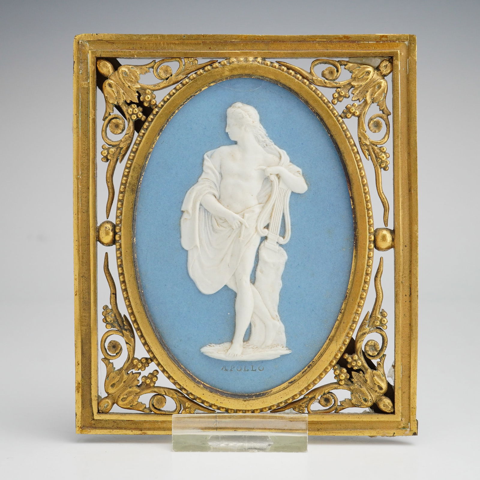 Wedgwood Light Blue Jasperware Plaque with Gilded Frame: This 19th-century Wedgwood light blue jasperware oval plaque features a white bas-relief figure of Apollo standing with a lyre, with the name APOLLO impressed at the base. The plaque is set within an