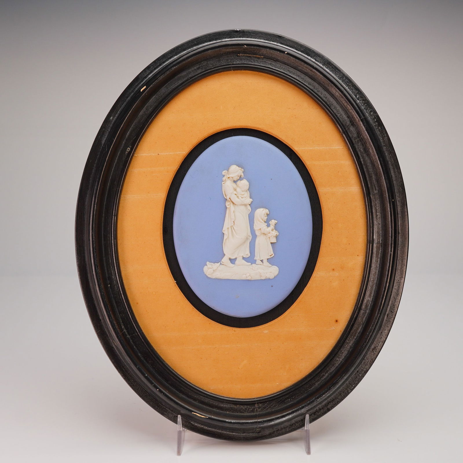 Framed Wedgwood Blue Jasperware Plaque: A framed oval ceramic plaque. The plaque features a white bas-relief cameo set against a pale blue ground. The relief depicts a standing mother holding an infant, accompanied by a smaller child who ca