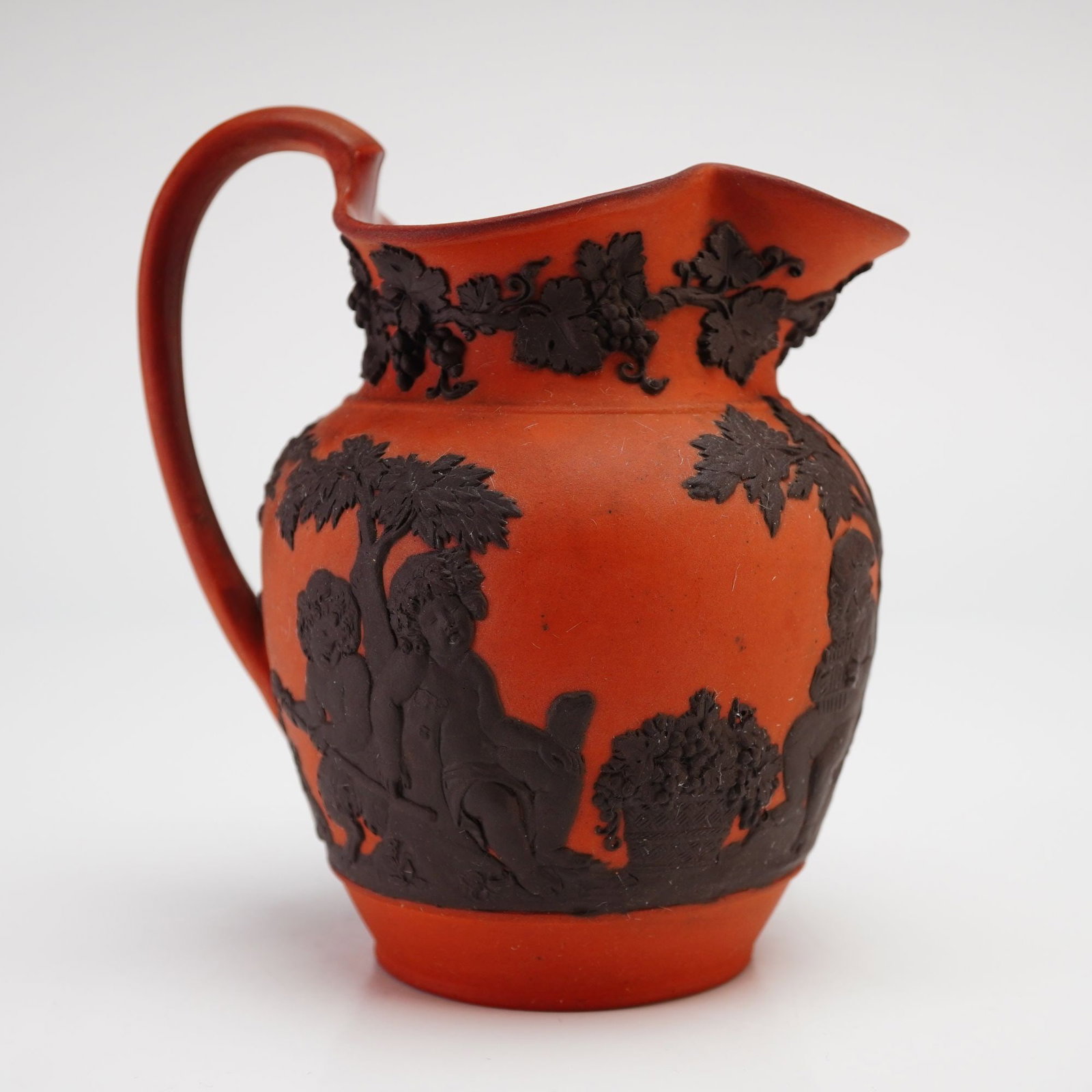 Wedgwood Rosso Antico Bacchanalian Boys Playing Pitcher: A Wedgwood Rosso Antico Bacchanalian Boys Playing pitcher featuring a terracotta-toned unglazed stoneware body. The vessel is decorated with black basalt sprigged-on relief motifs, including a grapevi