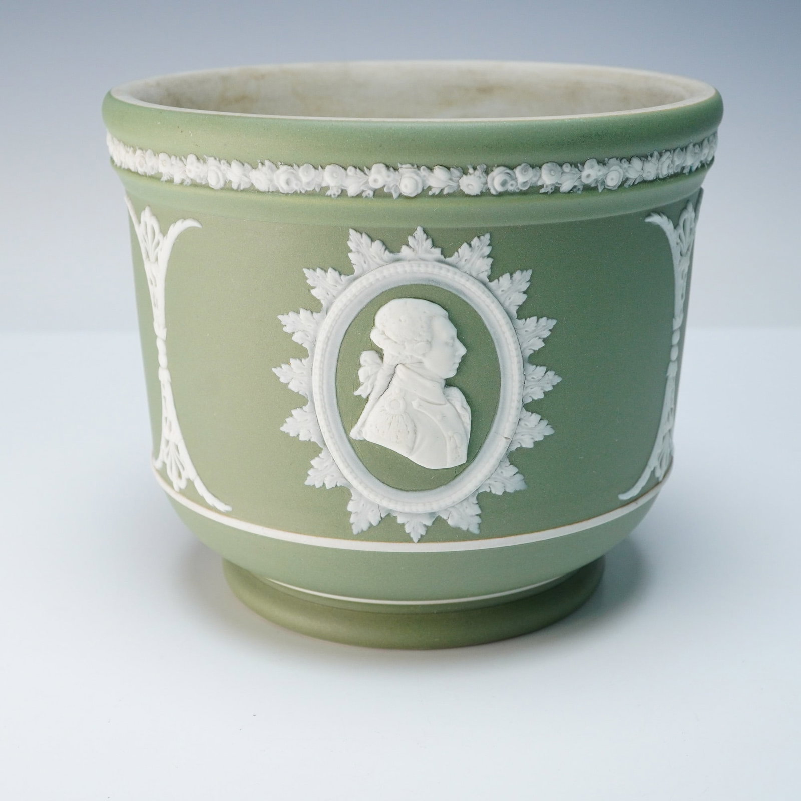 A Wedgwood Green Jasperware Jardiniere Founding Fathers Series: A Wedgwood green jasperware Jardiniere from the Founding Fathers Series, executed in a neoclassical style, featuring a matte sage green finish and white applied relief decoration. The ornamentation in