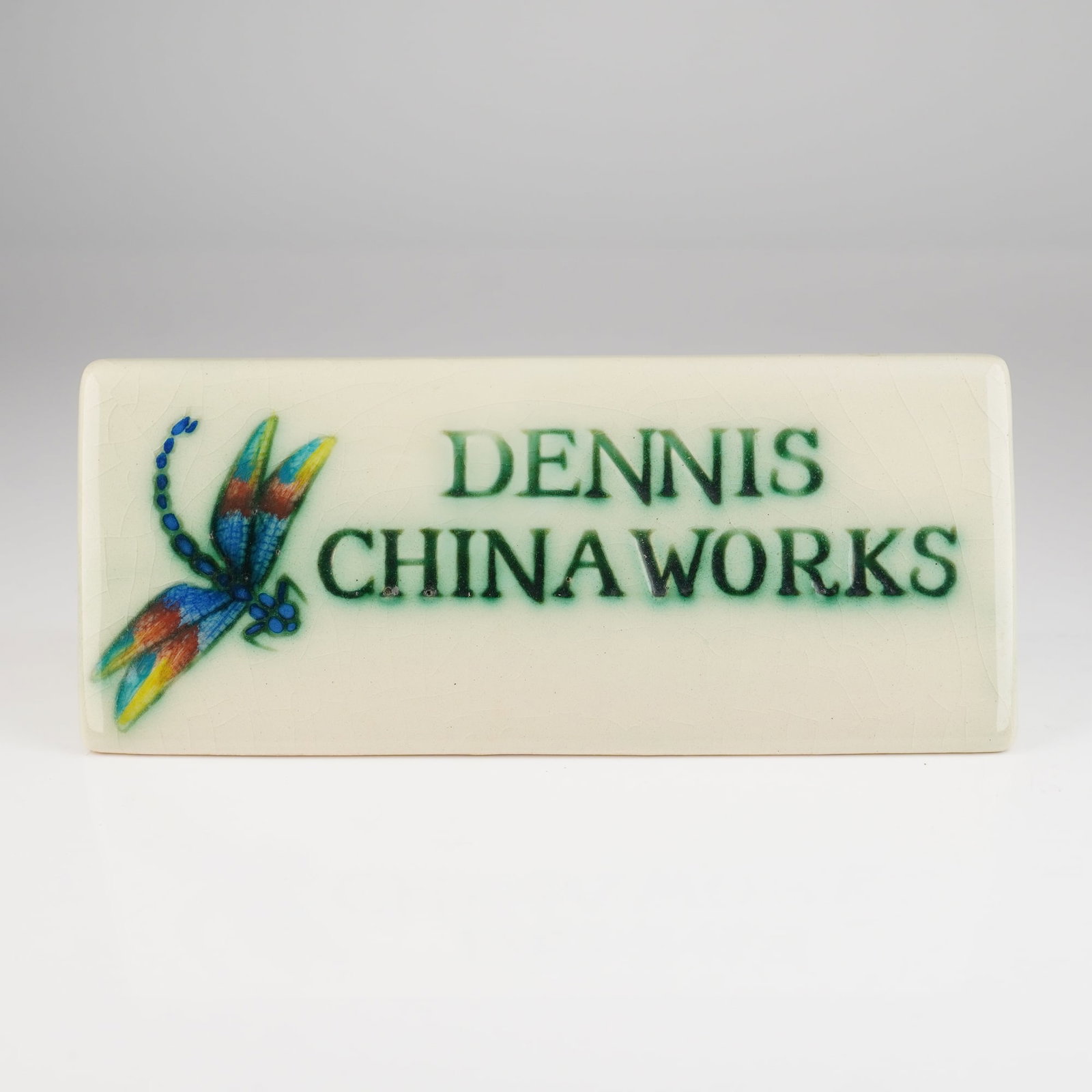 Dennis Chinaworks Ceramic Display Sign with Dragonfly Motif Dated 2001: A single ceramic counter plaque or display sign produced by Dennis Chinaworks. The piece is fashioned in a hollow, trapezoidal prism form from earthenware, finished with a pale cream or celadon glaze
