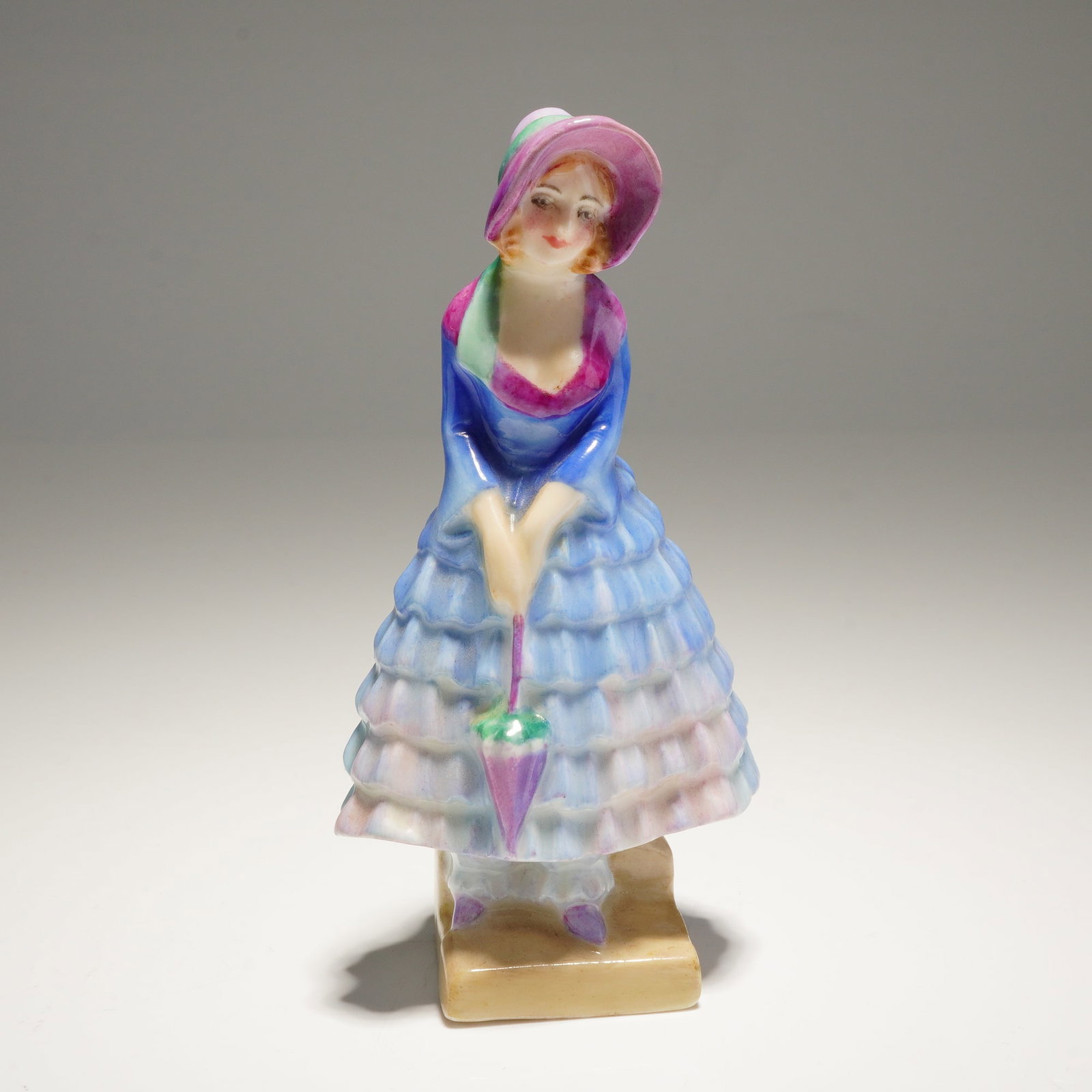Royal Doulton Miniature Figurine Priscilla M14: A single Priscilla M14 Royal Doulton Figurine, depicting a woman in a voluminous blue, pink, and lavender ruffled dress and large pink hat, marked on the base with the Royal Doulton logo. Dimensions2.