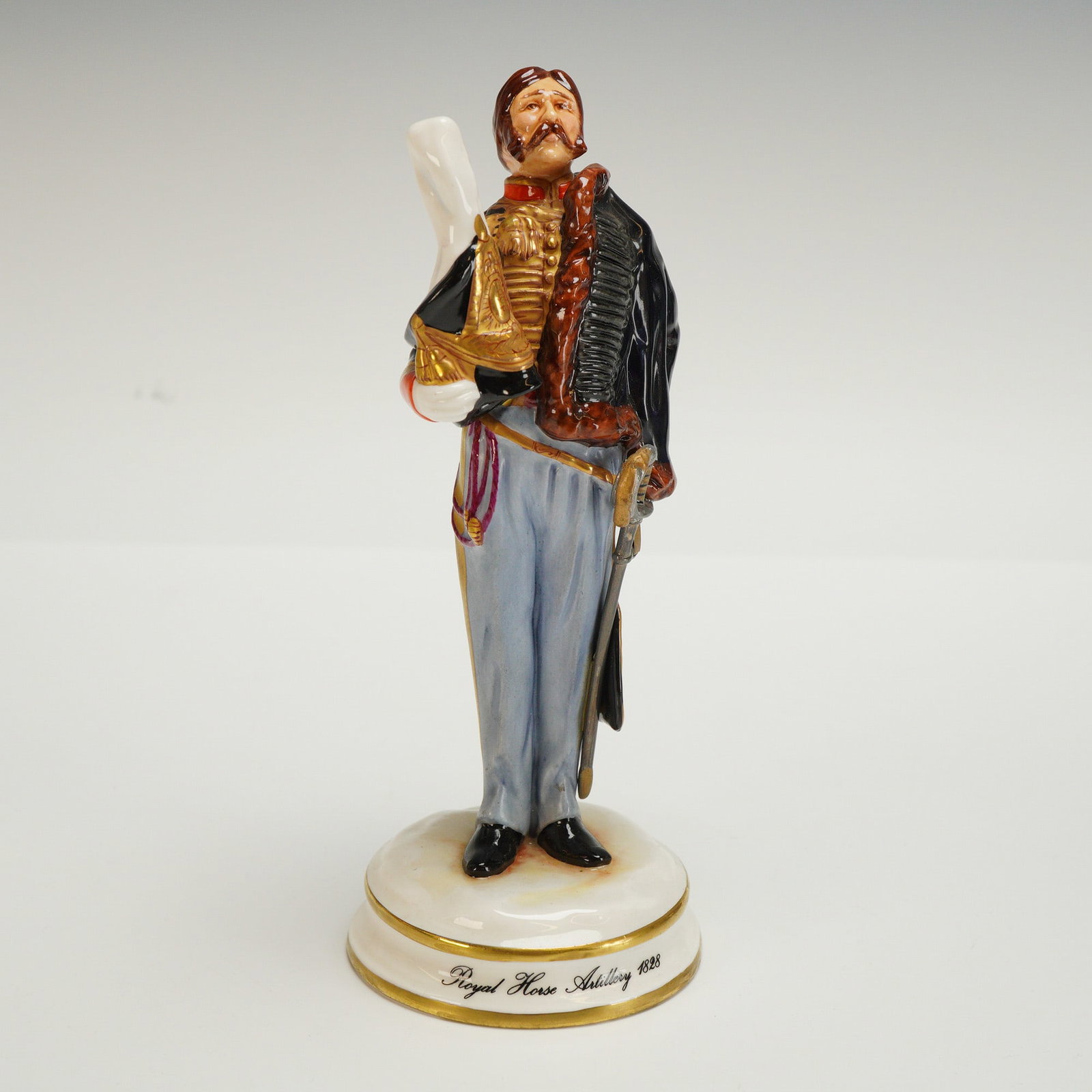 Michael Sutty Figurine Royal Horse Artillery 1828: Presented is a porcelain figure modeled as a Royal Horse Artillery Officer from 1828. The officer wears a traditional uniform with gilt detailing, with a coat draped over one arm and holding his white