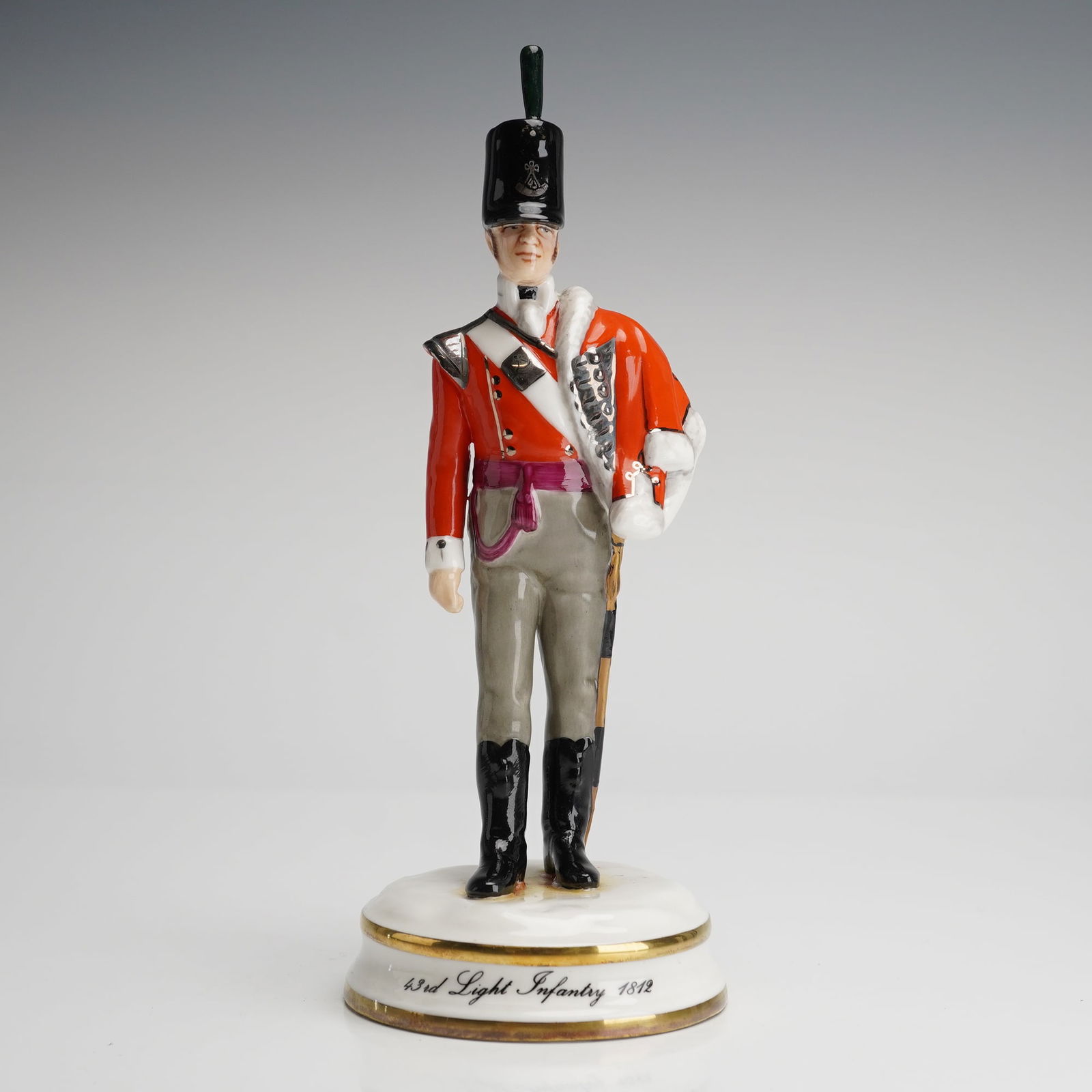 Michael Sutty Porcelain Figure 43rd Light Infantry 1812: A single glazed and hand-painted porcelain figurine by Michael Sutty, depicting a soldier of the 43rd Light Infantry from 1812. The figure is modeled wearing a red coat, grey trousers, and a tall blac