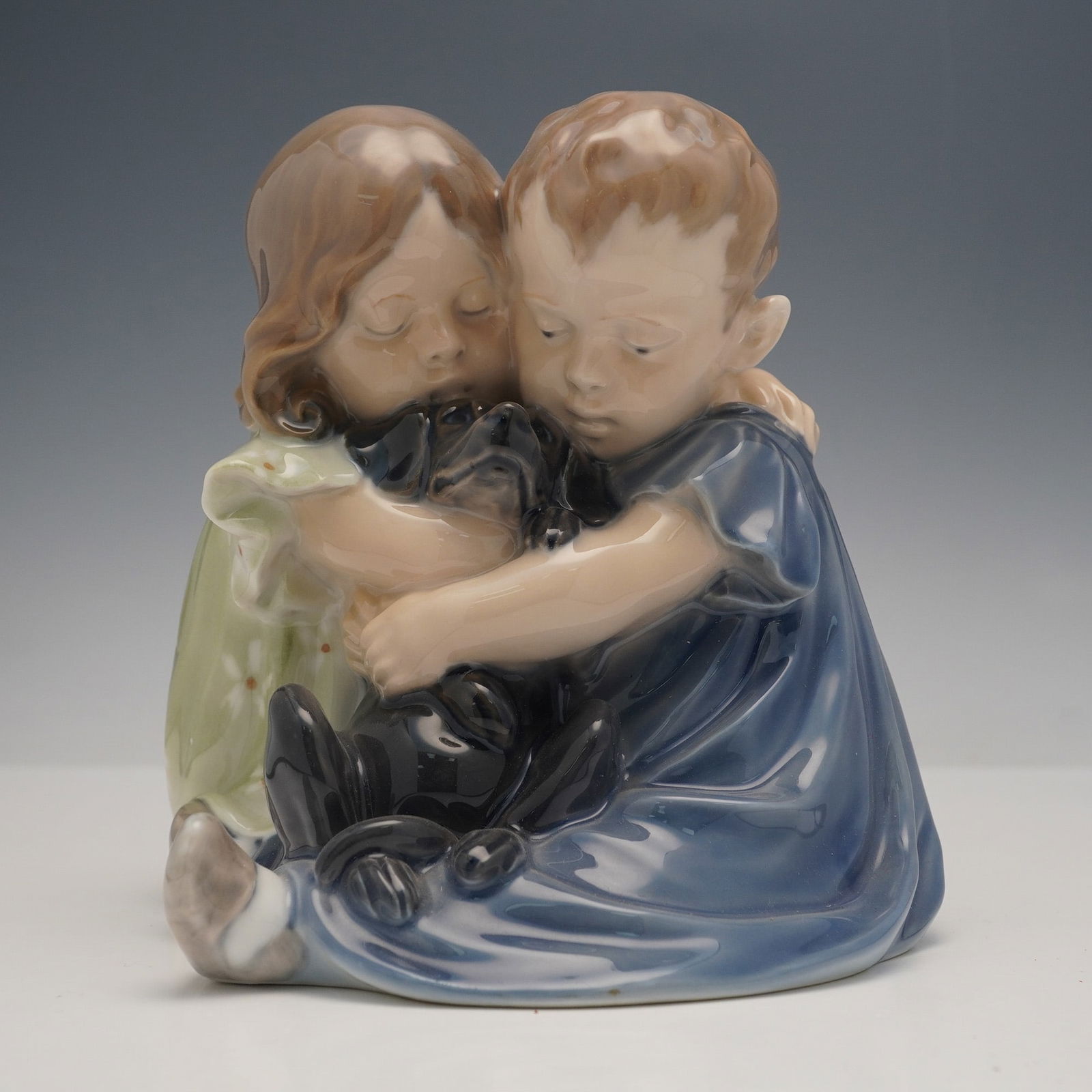 Royal Copenhagen Children with Dog Figurine: The piece depicts two young children, a boy and a girl, seated and embracing a small black puppy. The girl is rendered in a light green dress with white floral motifs, while the boy is dressed in blue