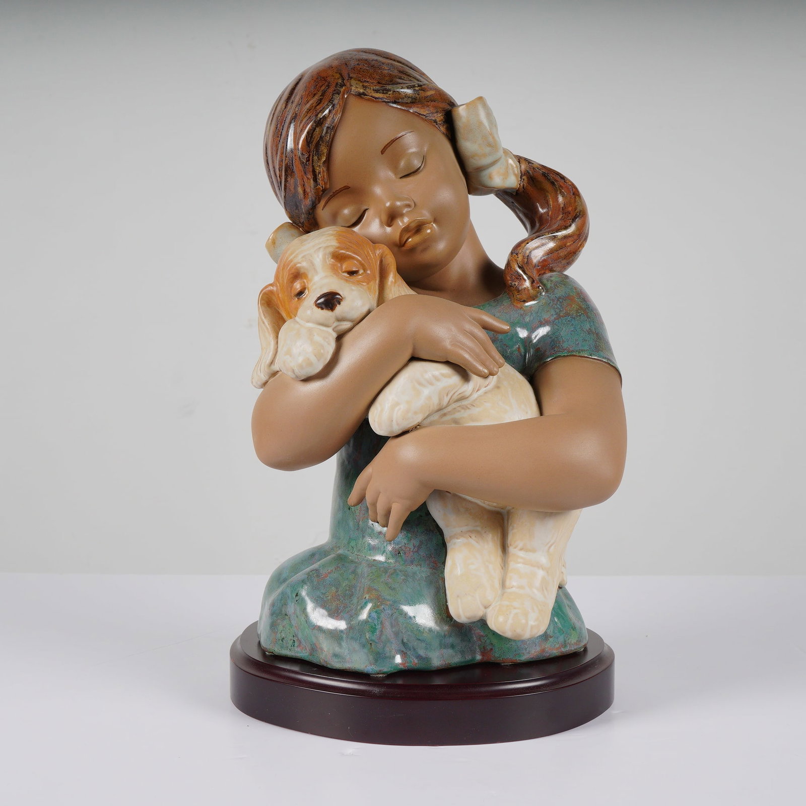 Lladro Gabriela Gres Porcelain Figurine 1012355 with Wood Base: A gres porcelain figurine of a girl with brown skin hugging a tan and white dog, featuring a Lladro backstamp and including a circular wood base. Dimensions8.50"L x 7.00"W x 13.00"H