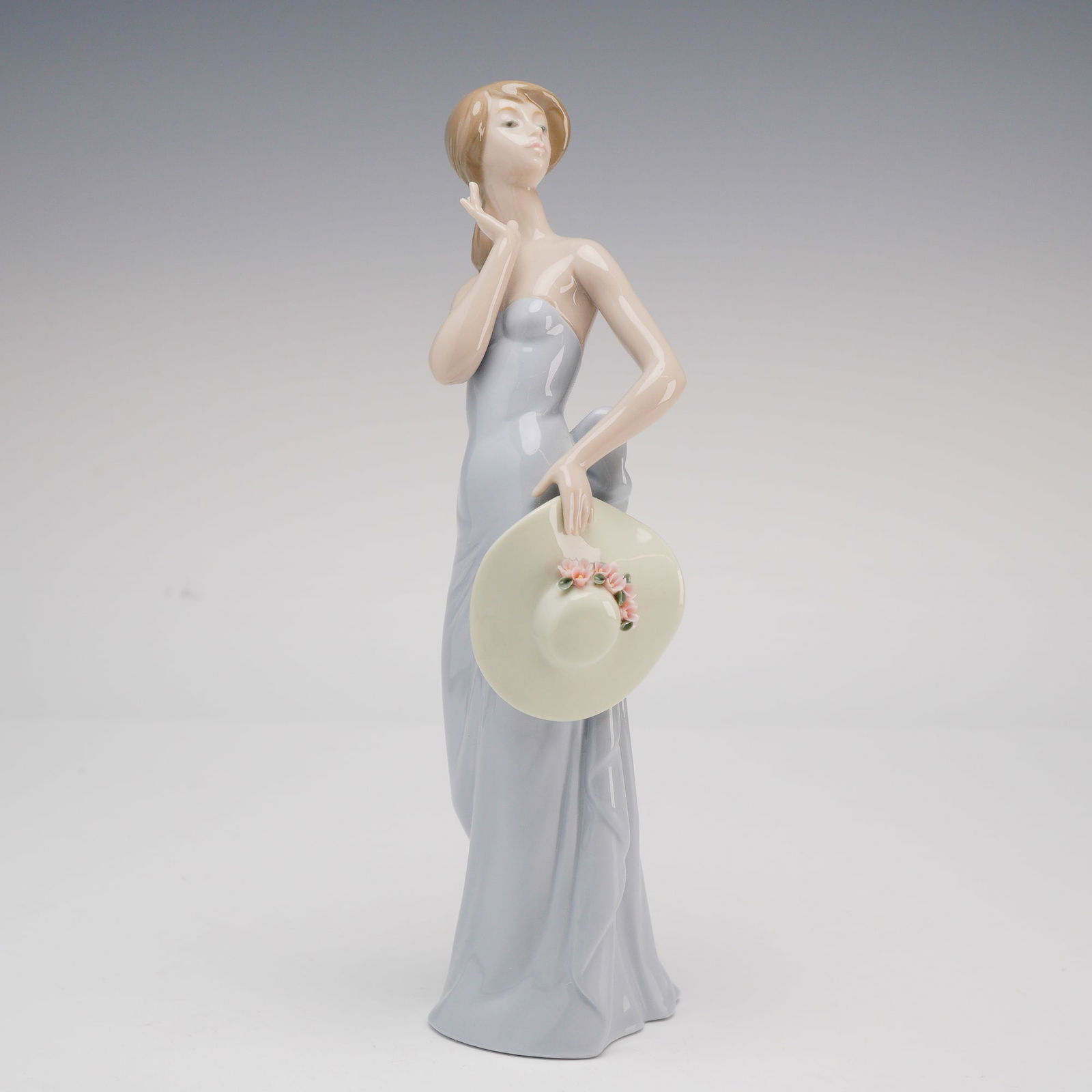 Lladro Coquette Glazed Porcelain Figurine No. 5599: A fine glazed porcelain study titled "Coquette" (Dama Esquiva), model number 5599, produced by Lladro. Designed by sculptor Juan Huerta and issued in 1989, this elegant figurine depicts a young woman