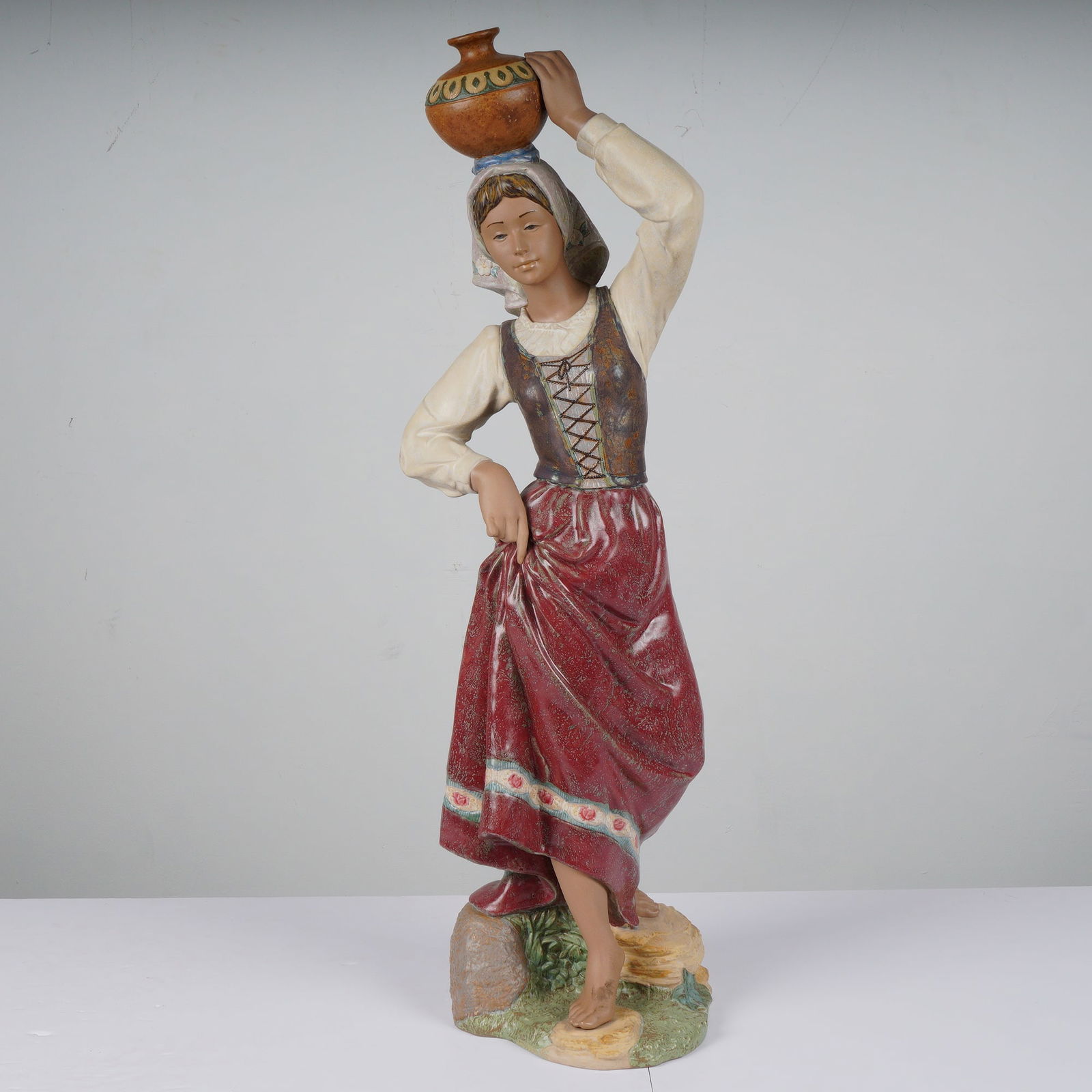 Lladro Returning From the Well Porcelain Figurine 1012342: A Lladro porcelain figurine titled Returning From the Well, model 1012342, depicting a woman in traditional dress carrying a jug on her head with a red, white, and brown finish, featuring a Lladro