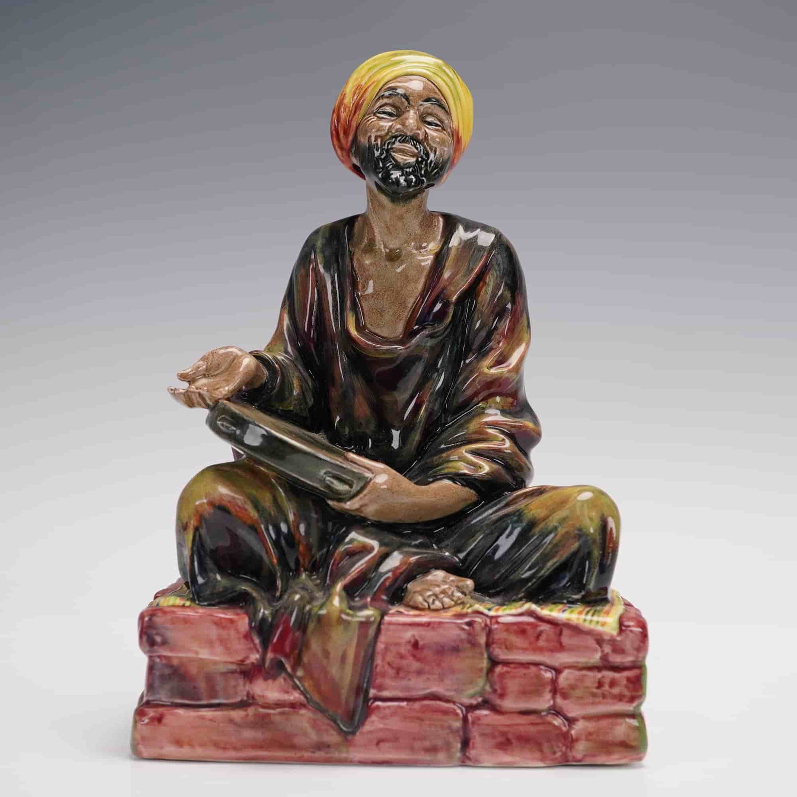 Royal Doulton Figurine, Mendicant HN1365: Charming and richly colored Royal Doulton figurine titled, Mendicant, HN1365. Designed by Leslie Harradine, this figure depicts a joyful seated man in flowing robes, with a bright yellow and red turba